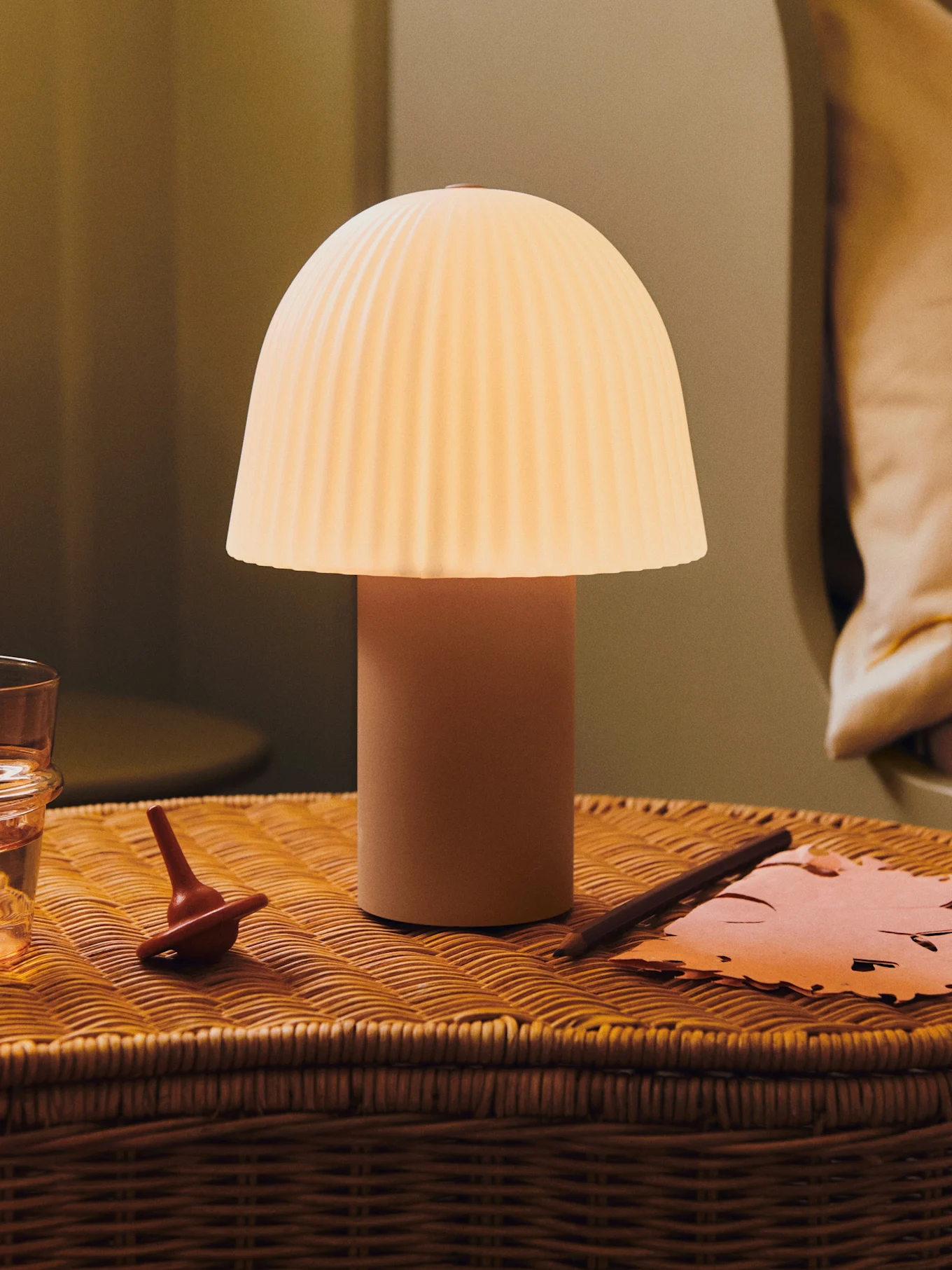 Une lampe champignon lumineuse avec un abat-jour plissé sur une table de chevet en osier avec un verre d'eau, une toupie et du papier.
