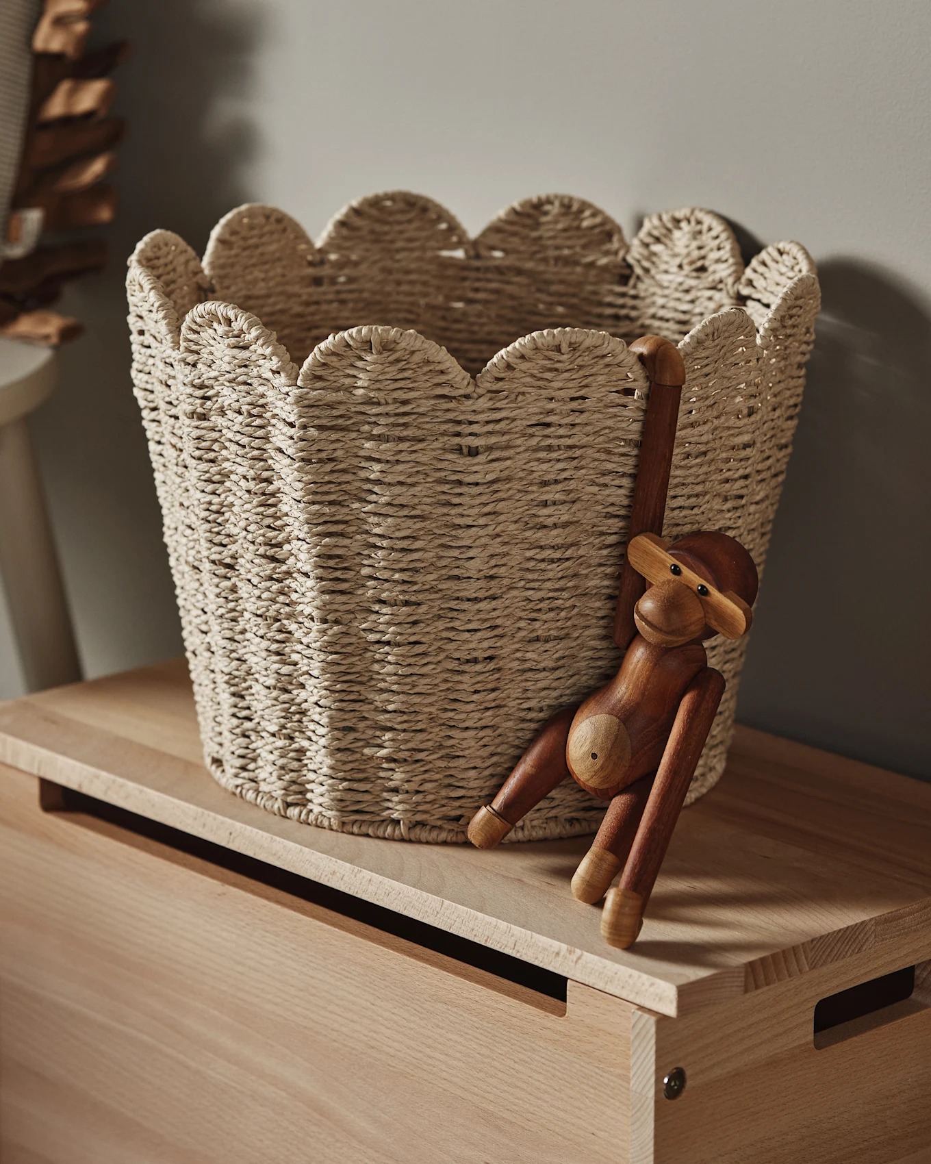 Un panier tressé clair avec un bord festonné posé sur une surface en bois, avec une figurine de singe en bois suspendue sur le côté.
