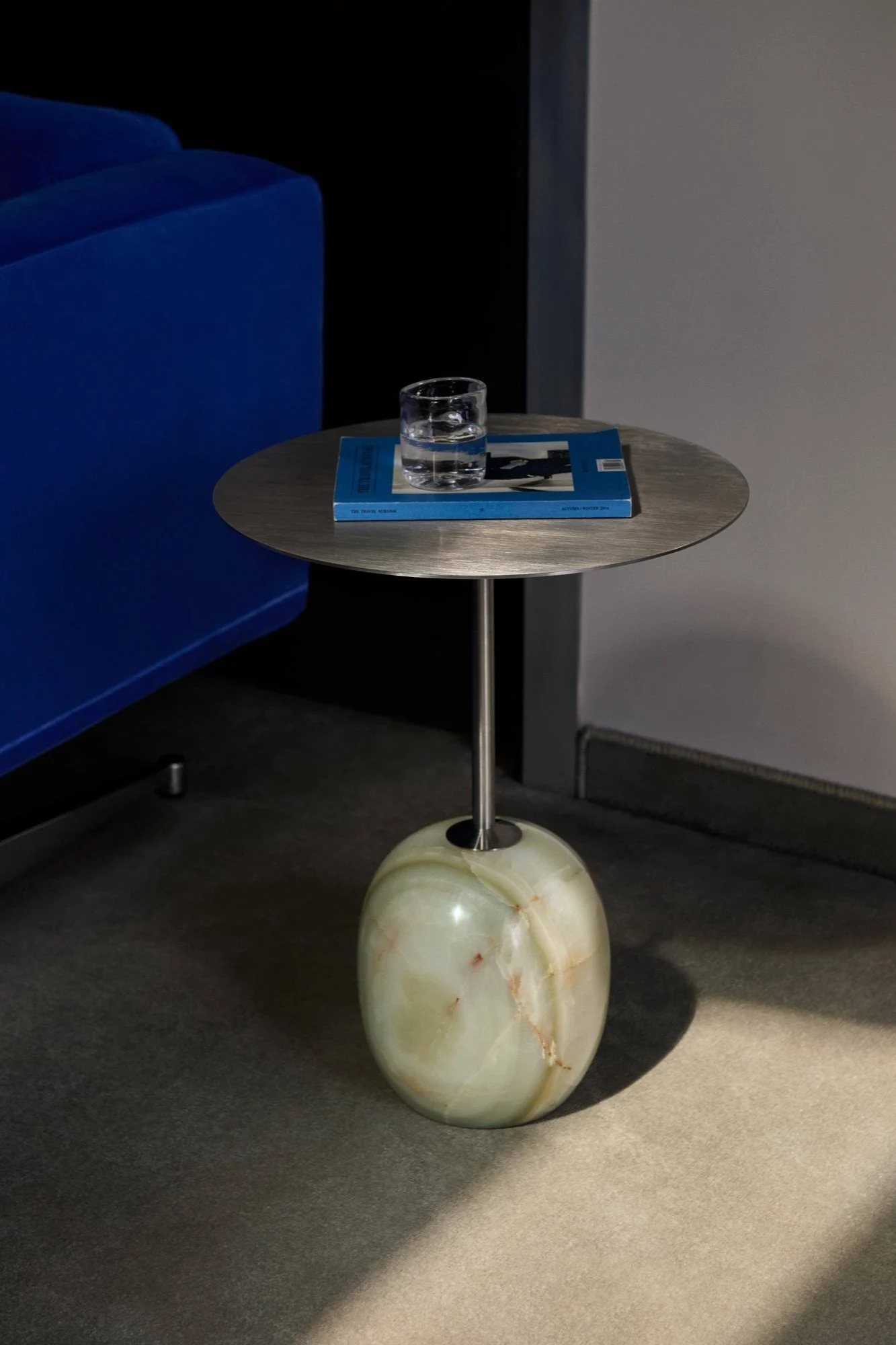 Table d'appoint avec plateau en métal brossé, piètement rond en onyx vert, verre à eau, hêtre bleu, à côté d'un canapé bleu.
