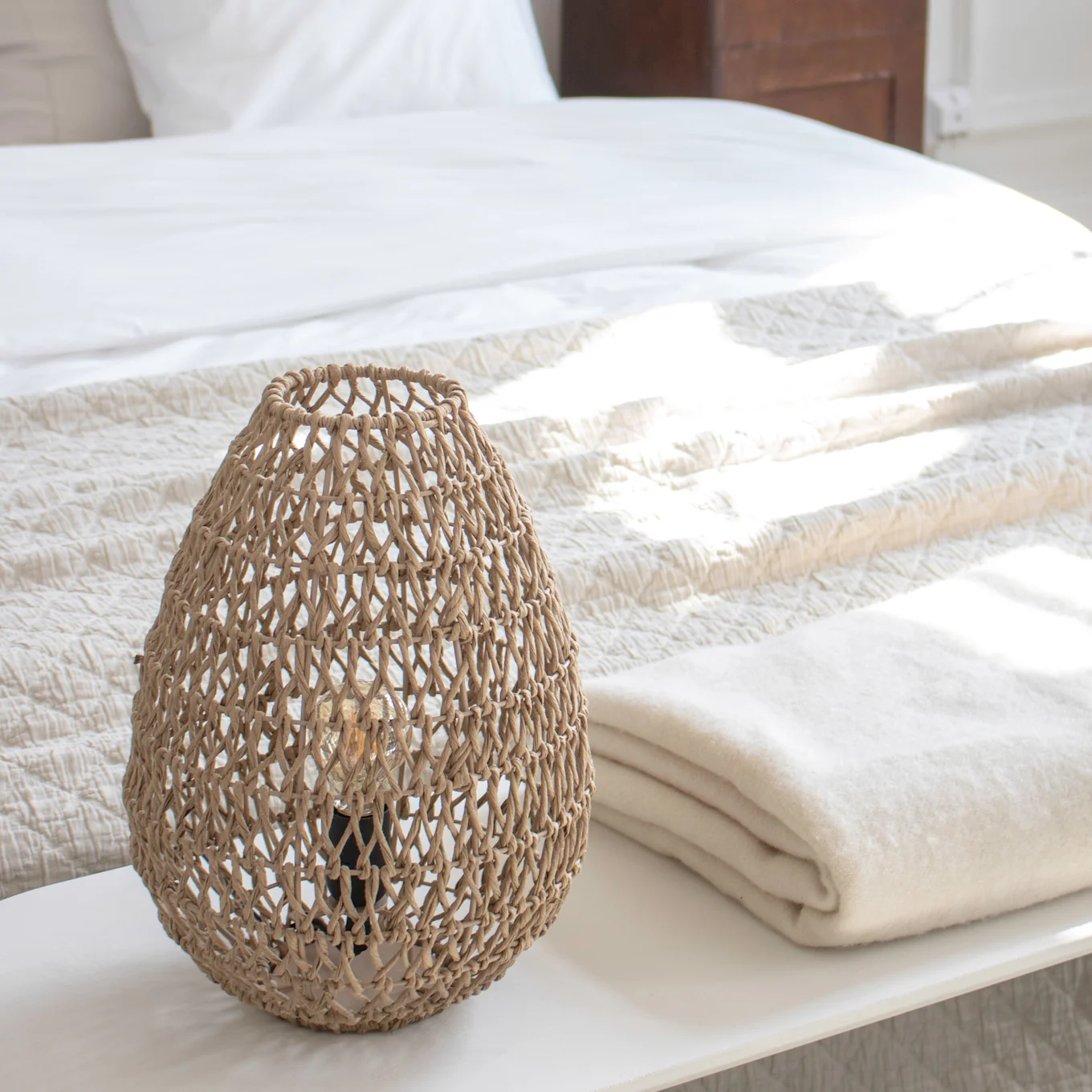 Lampe tressée et couverture pliée sur un banc dans une chambre lumineuse.