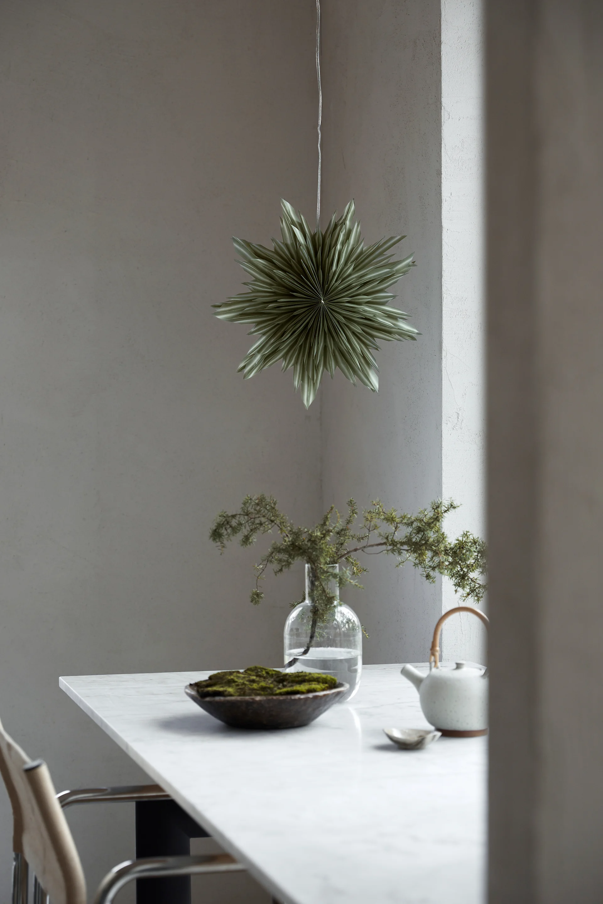 La lanterne en papier Helena, de couleur verte, est suspendue à la fenêtre au-dessus d'une plante verte en pot. 