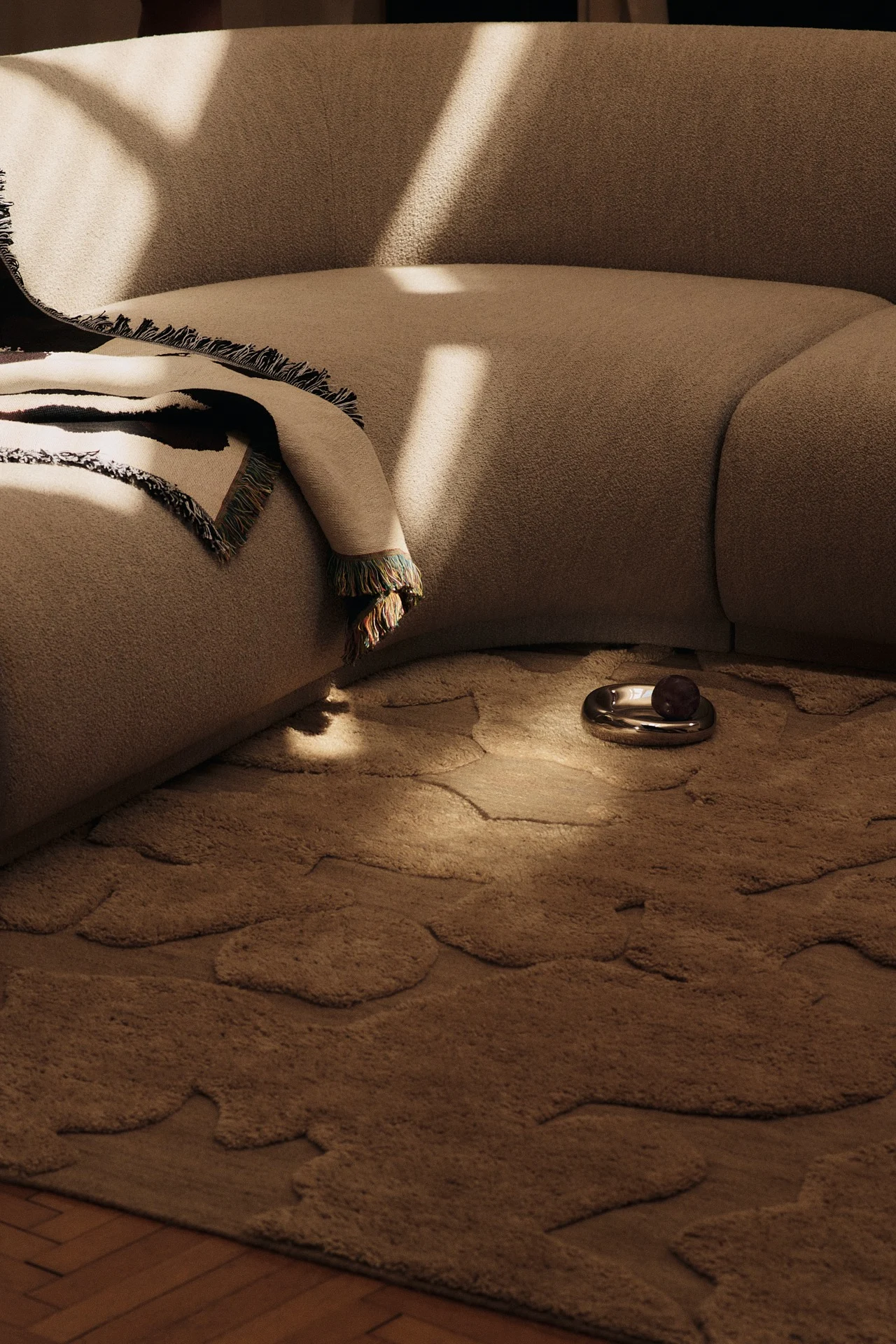 Canapé beige incurvé avec plaid à motifs, tapis beige aux formes organiques et plat argenté avec fruit foncé dans une pièce ensoleillée.