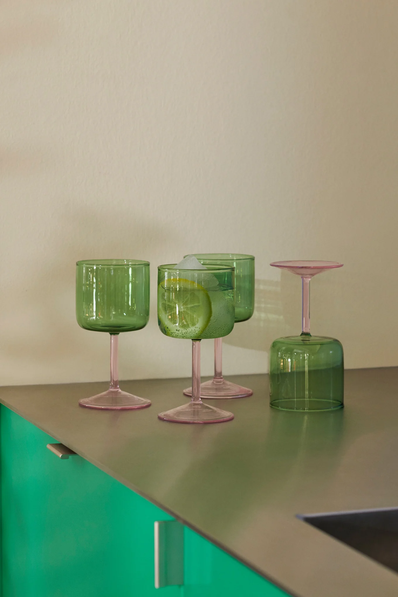 Quatre verres verts aux pieds roses posés sur un plan de travail métallique. L'un des verres contient une boisson au citron et des glaçons ; un autre est à l'envers.