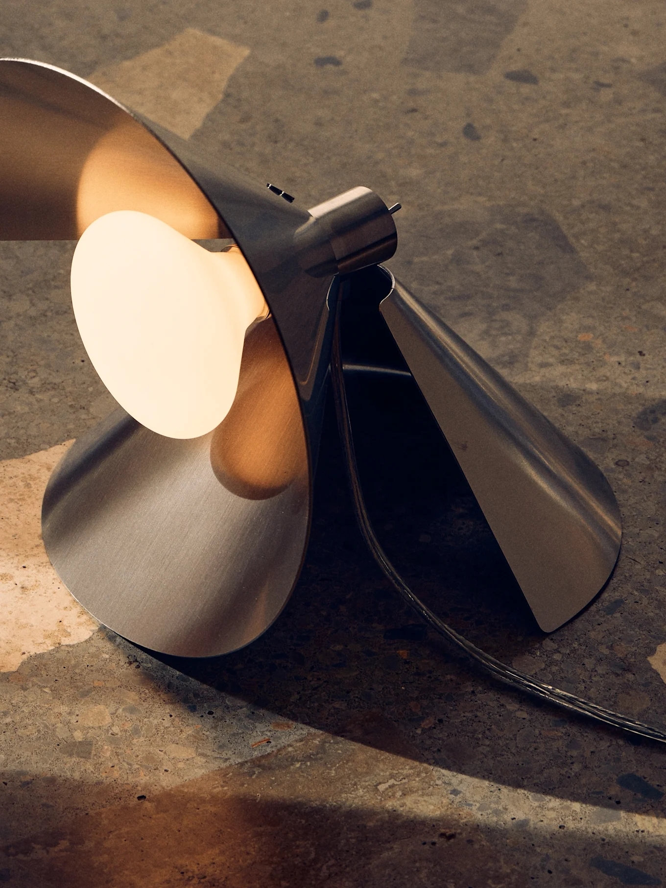 Lampe de table moderne en métal brossé, design sculptural plié et ampoule chaude exposée sur une surface texturée.
