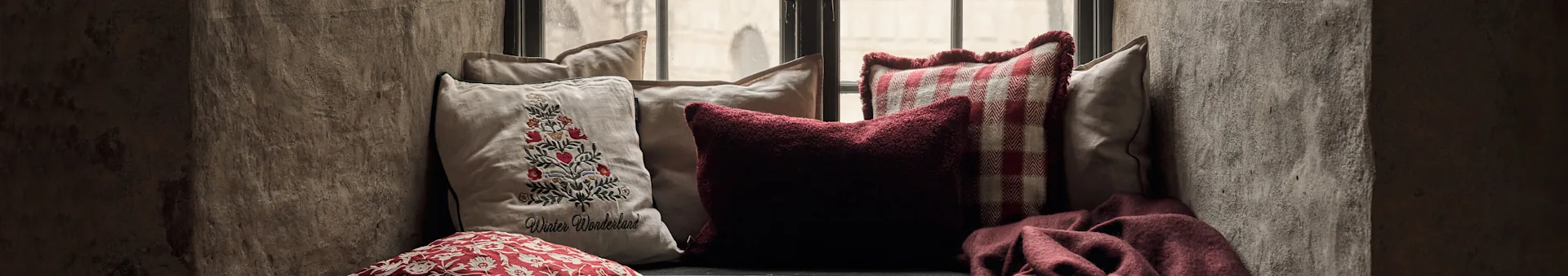 Banquette de fenêtre douillette avec coussins: un coussin brodé sapin de Noël, un rouge texturé, un à carreaux rouges et un plaid rouge.