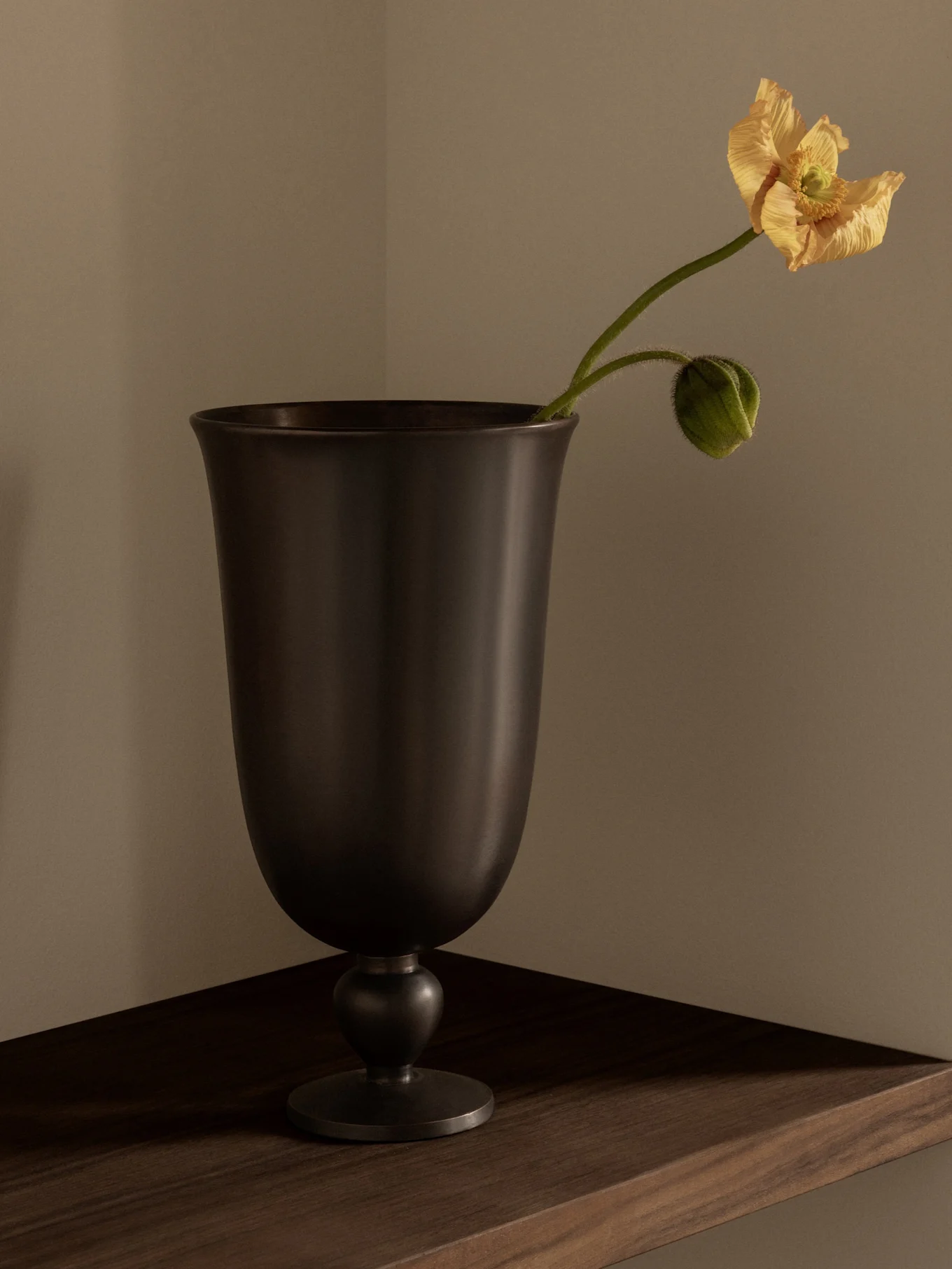 Un vase sombre en forme de calice contenant un délicat coquelicot jaune-orange et un bouton vert sur une étagère en bois.