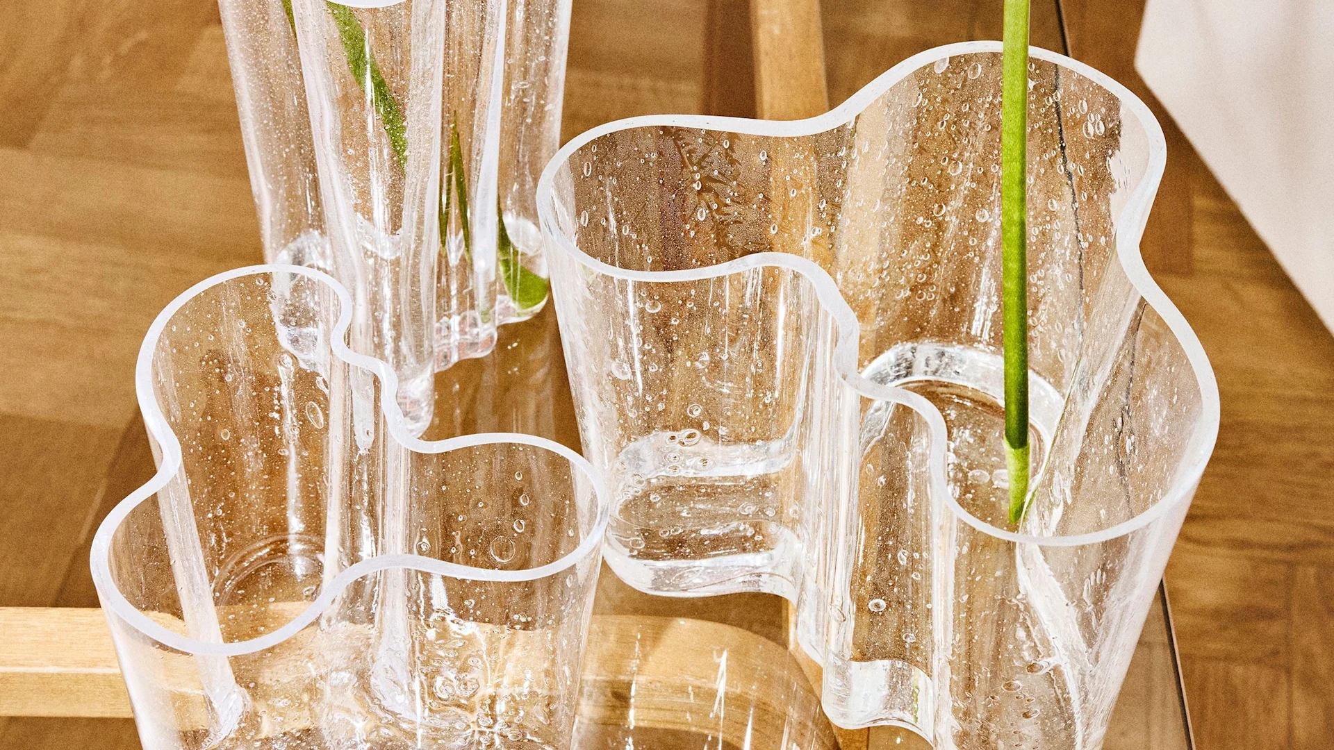 Plusieurs vases en verre transparent aux formes ondulées organiques et bulles d'air intégrées, contenant des tiges vertes, sur une surface en bois.