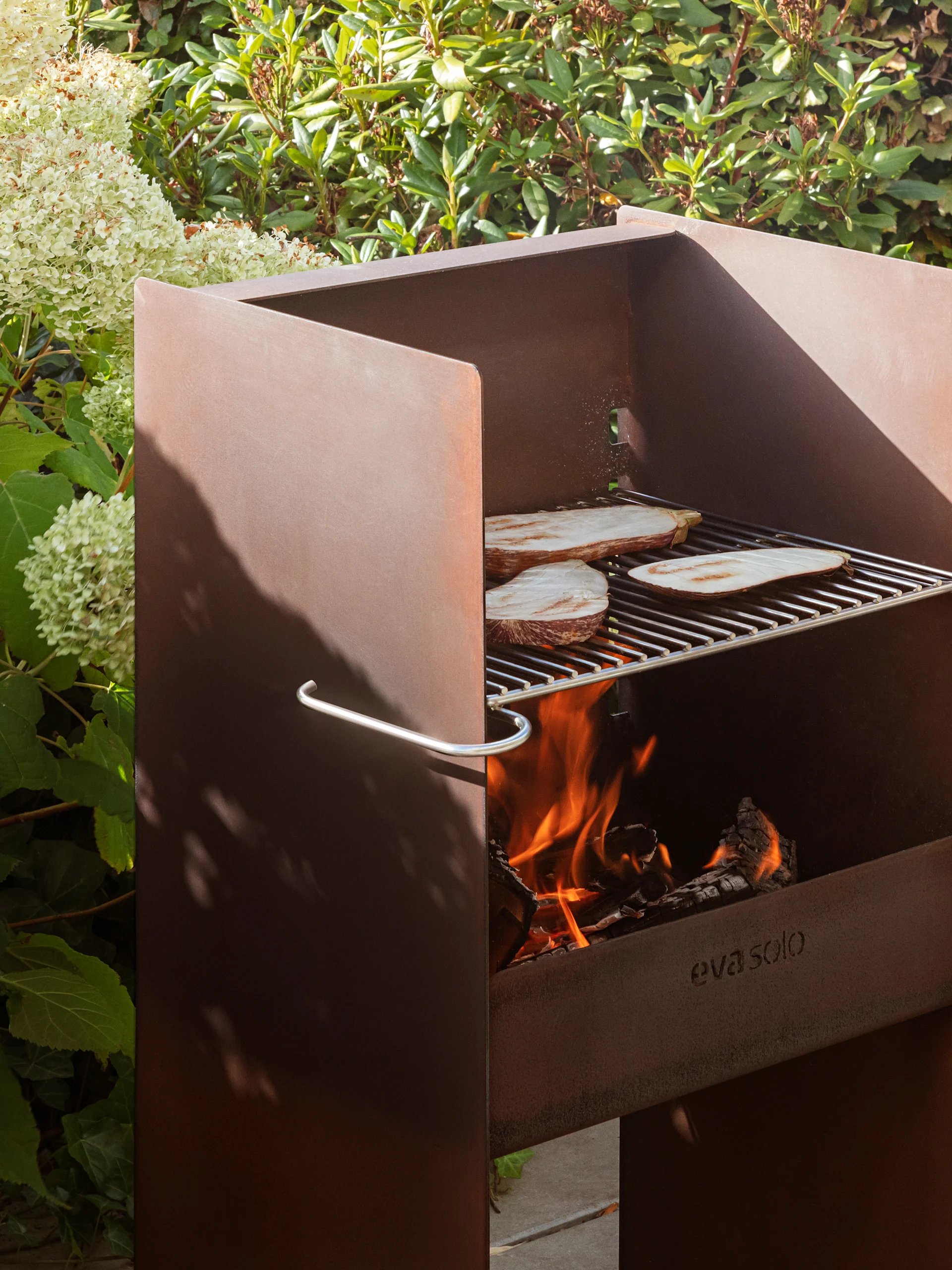 Grill extérieur en métal rouillé Eva Solo avec aubergines sur la grille, feu ardent et feuillage luxuriant.