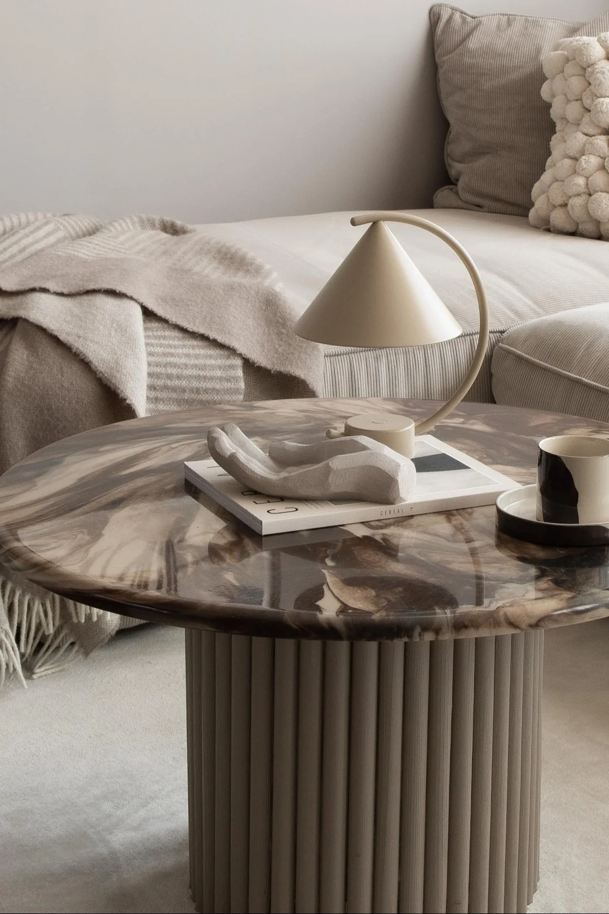 Une table basse avec plateau en marbre est décorée d'une tasse à café, d'une main sculpturale et de la lampe de table Meridian de Ferm Living.