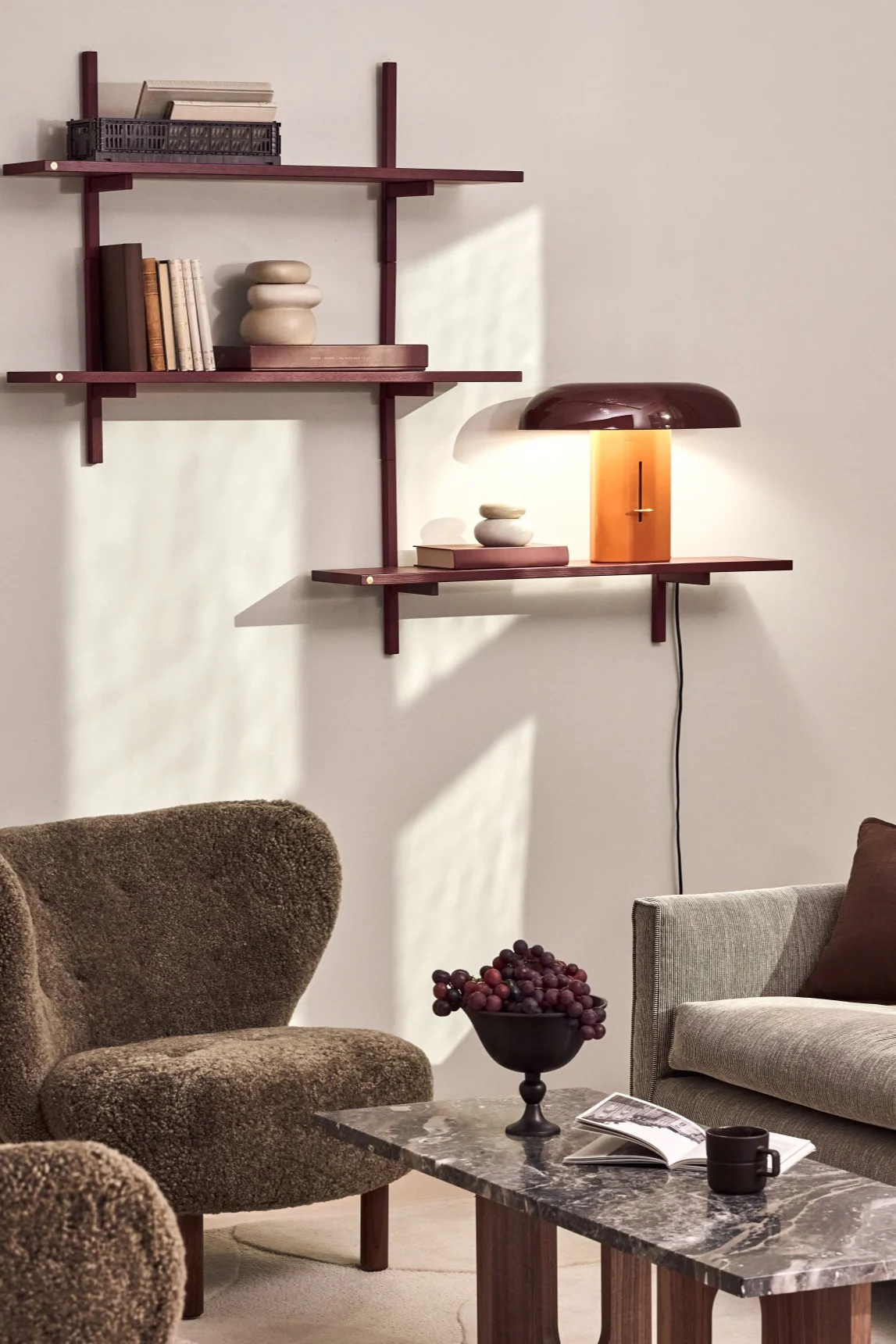 Un salon chaleureux avec des étagères murales brun rougeâtre, une lampe à abat-jour bombé et base orange, un fauteuil marron texturé et une table basse en marbre avec des raisins et un livre.