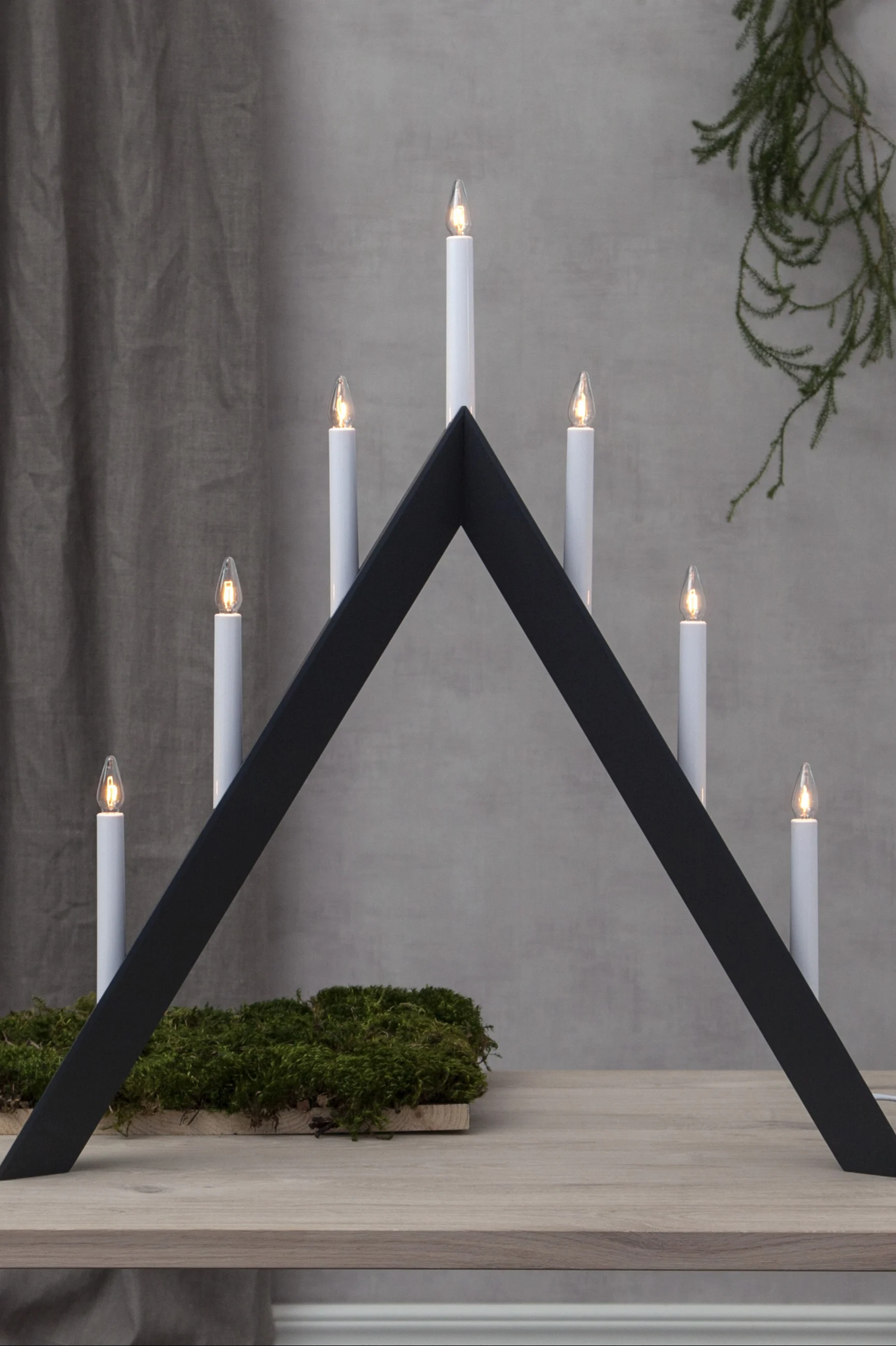 Un chandelier triangulaire blanc avec sept bougies électriques allumées sur une surface blanche, avec un sapin de Noël flou en arrière-plan.