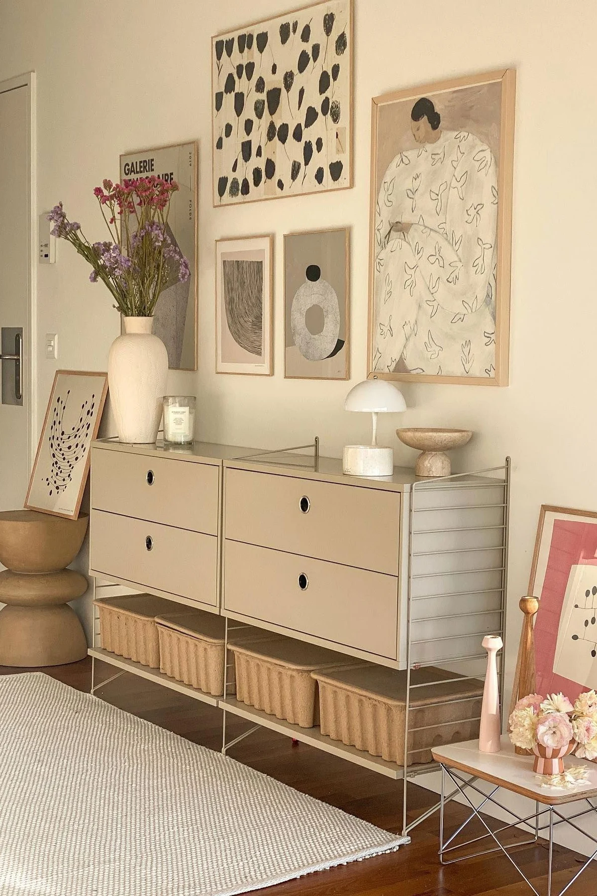 Une pièce moderne avec un meuble modulaire beige, des tiroirs et des paniers de rangement. Un vase de fleurs, une lampe et de l'art abstrait ornent l'espace.