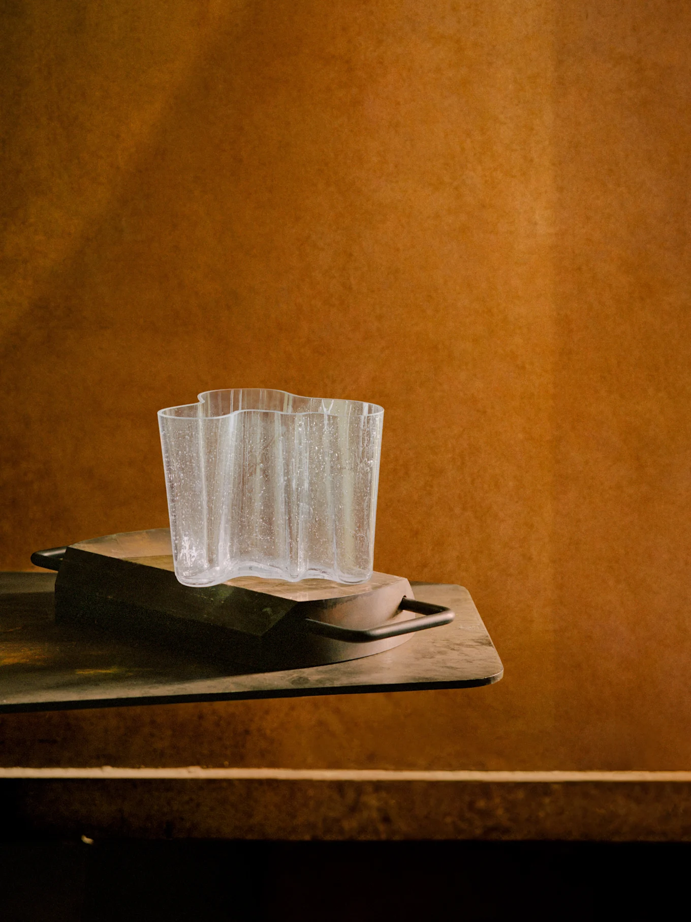 Vase en verre clair ondulé avec des gouttelettes d'eau sur un plateau en bois foncé, devant un fond texturé marron chaud.