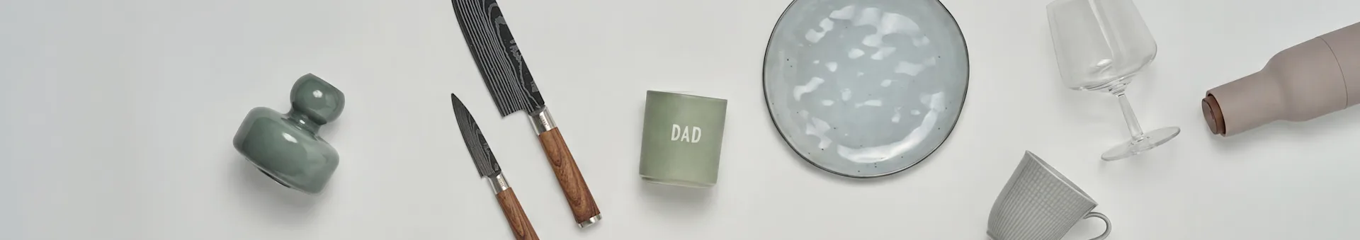 Une vue à plat d'articles de cuisine et de cadeaux : un vase vert, deux couteaux, une bougie 'DAD', une assiette en céramique grise, un verre à vin, une bouteille beige et une tasse grise.