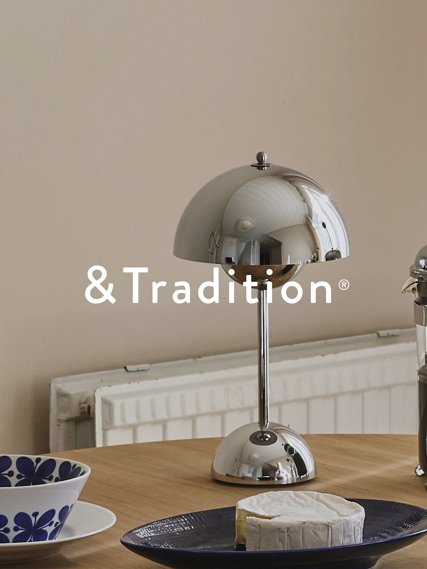 Une lampe de table argentée en forme de dôme, un fromage rond sur une assiette bleu foncé et un bol à motifs bleus sur une table en bois. Logo &Tradition.