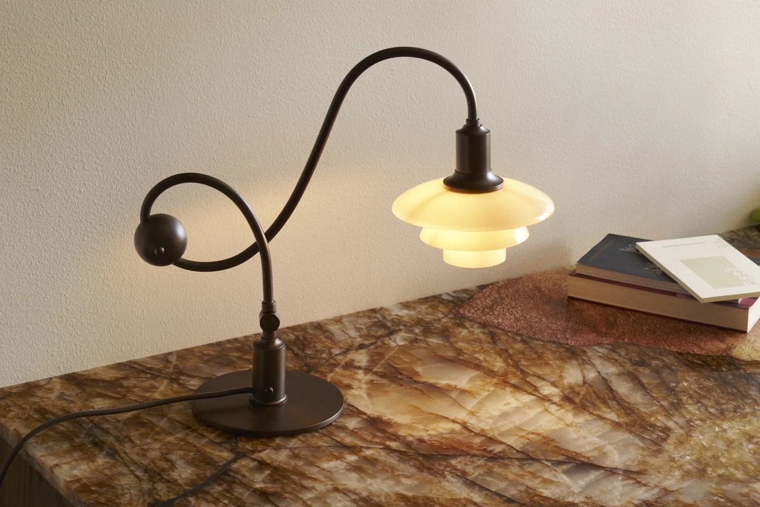 Lampe de table en métal foncé avec tige courbée et abat-jour blanc étagé et lumineux sur une surface marbrée avec des livres.