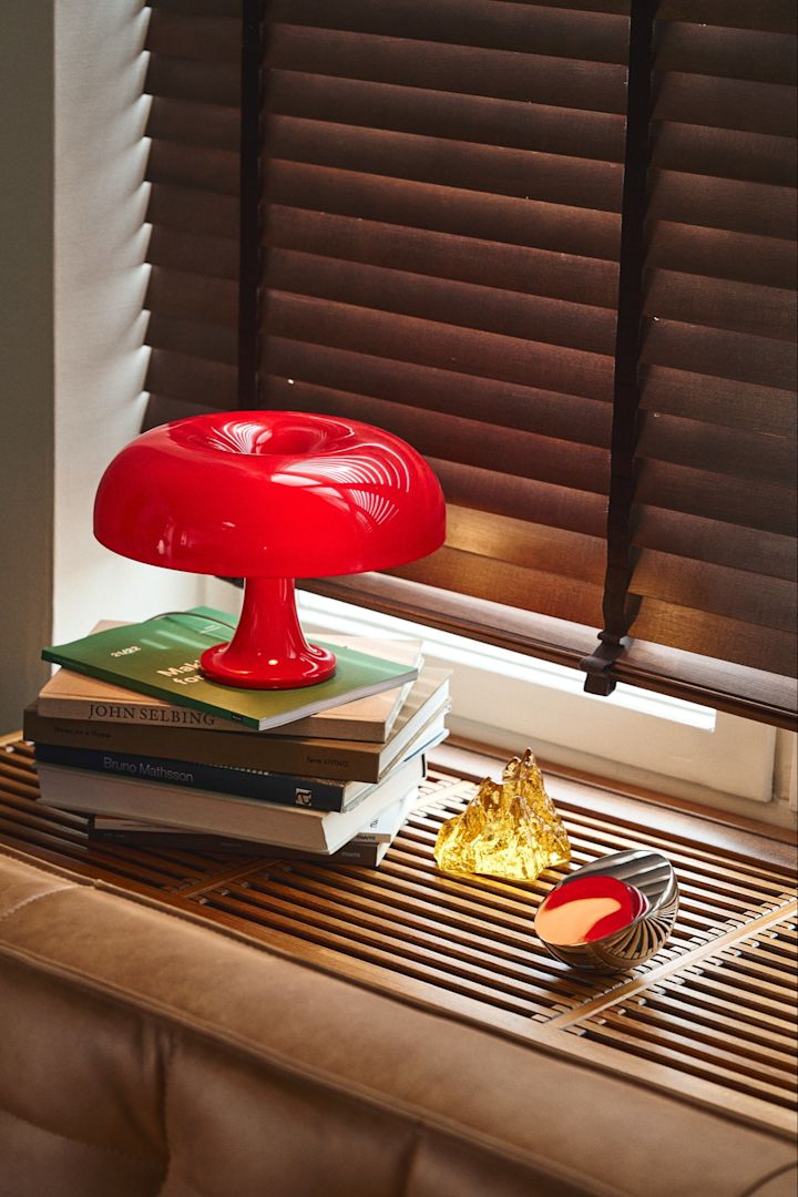 La lampe Nessino en forme de champignon rouge est posée sur une pile de livres sur un rebord de fenêtre. 