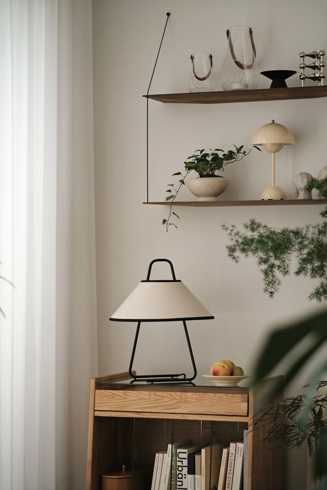 Salon moderne avec une lampe de table crème et noire sur une armoire en bois clair, des étagères décoratives et des plantes.