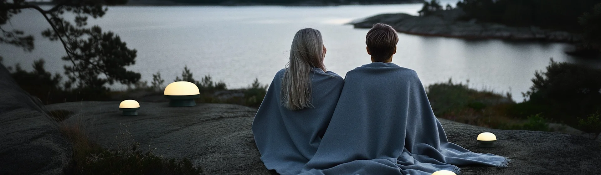 Deux personnes sous une couverture bleue assises sur des rochers, regardant un lac paisible avec des îles au crépuscule. Quatre lampes portables éclairent la scène.