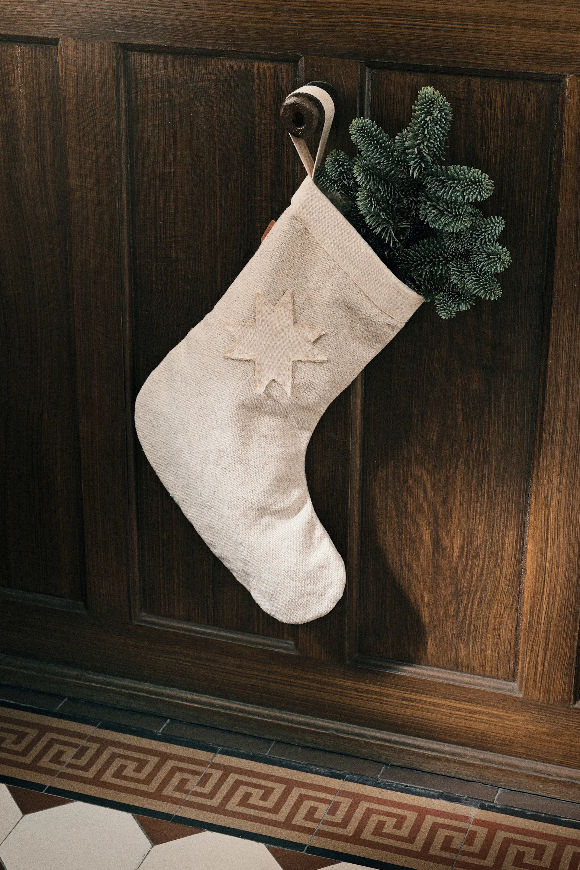 Fabriquer des décorations de Noël avec du sapin : voici la chaussette de Noël Vela de ferm LIVING, décorée de branches de sapin et suspendue à un crochet fixé au mur.