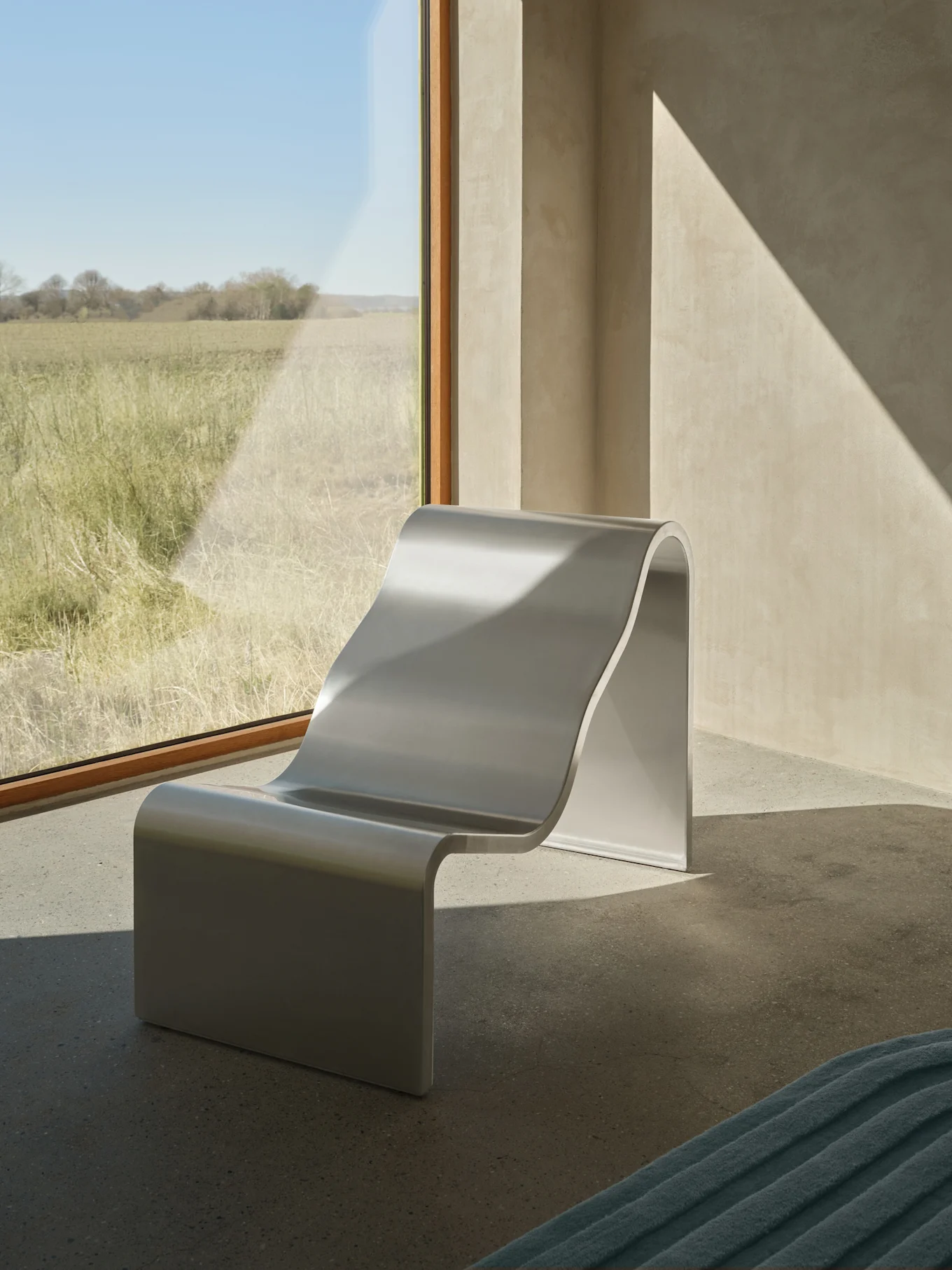Un fauteuil de salon moderne en forme de S argenté est placé près d'une grande fenêtre donnant sur un champ herbeux ensoleillé, avec des motifs de lumière diagonaux sur les murs.