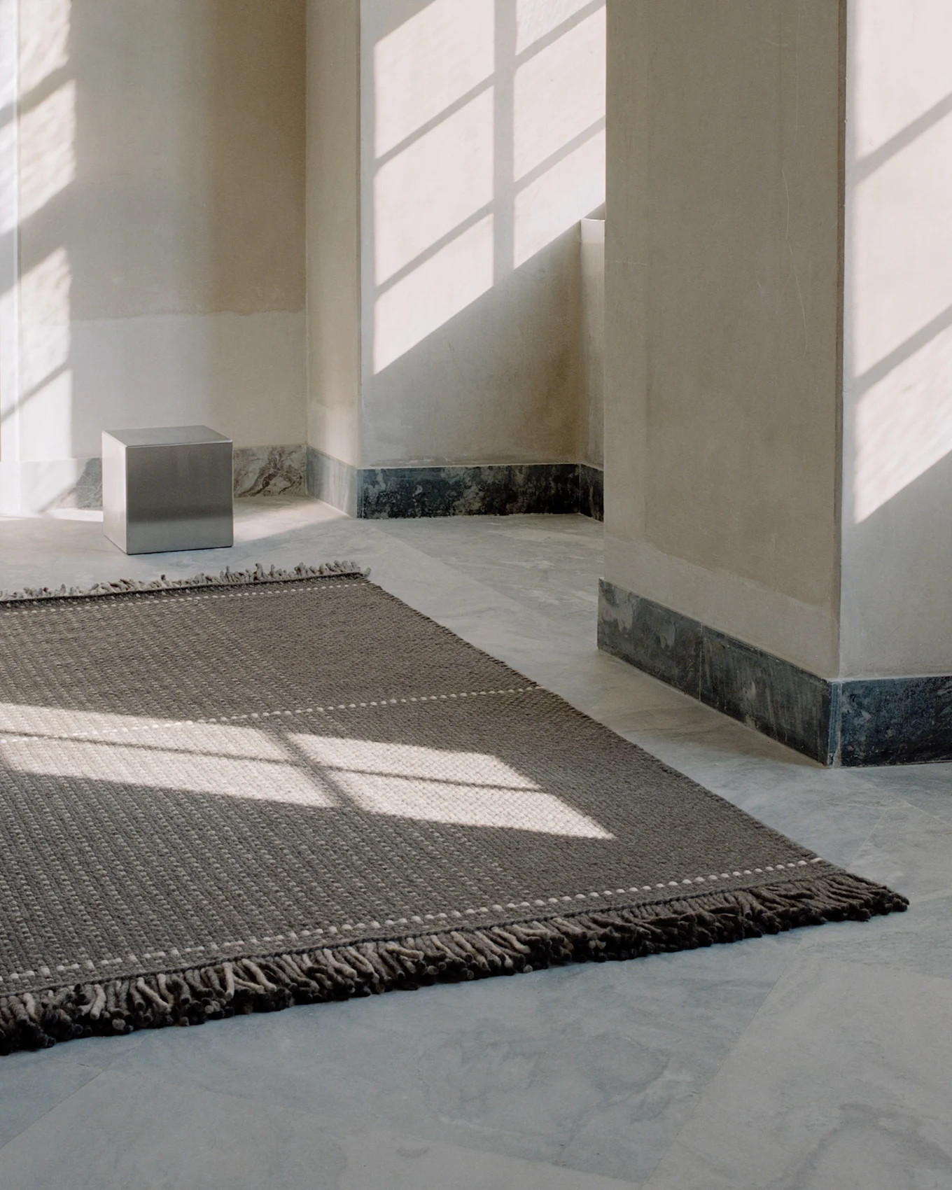 Un tapis texturé gris foncé avec des points clairs sur un sol carrelé clair, éclairé par des ombres de fenêtre dans une pièce minimaliste.
