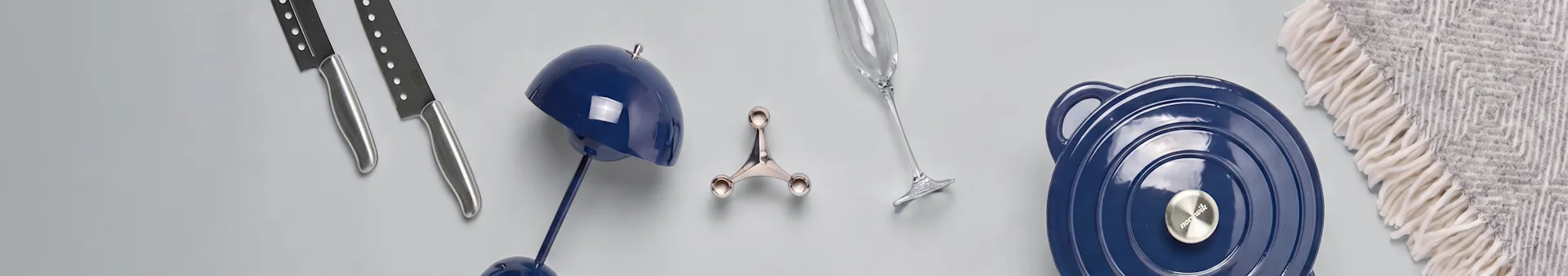 Assortiment d'ustensiles de cuisine et de décoration intérieure: deux couteaux argentés, une lampe décorative bleue, un outil de cuisine argenté, une flûte à champagne, une cocotte bleue avec couvercle et un plaid à motifs.
