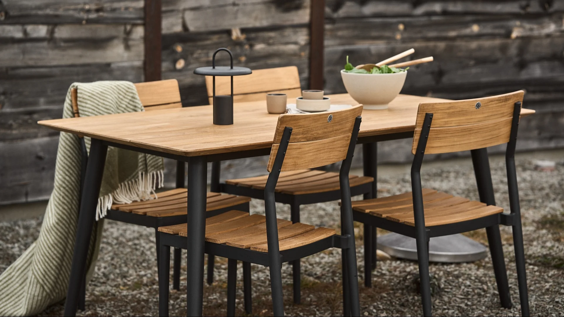 Ensemble de salle à manger d'extérieur avec une table et quatre chaises en bois, un plaid rayé, une lanterne et un saladier.