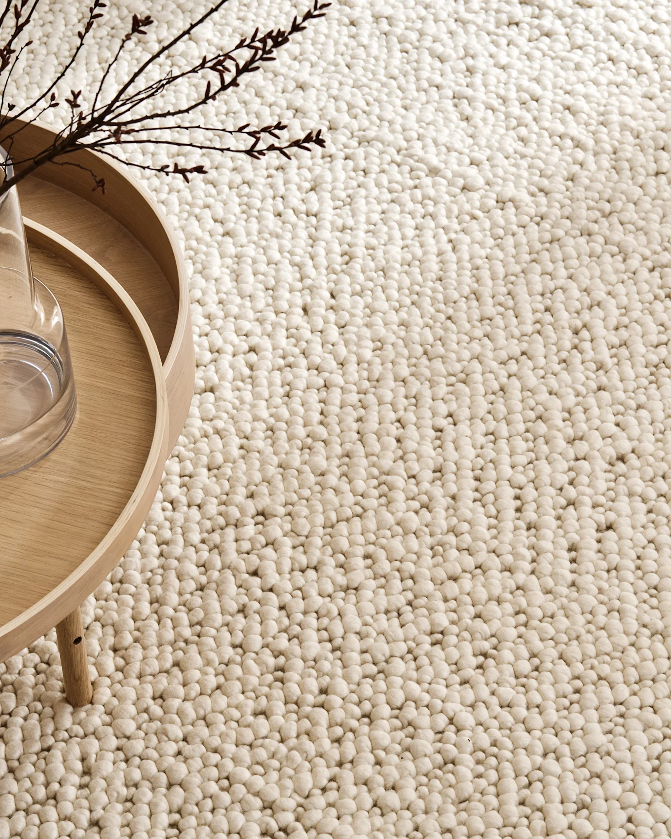 Tapis texturé beige clair avec une surface bosselée; une table en bois et des branches nues sont partiellement visibles.