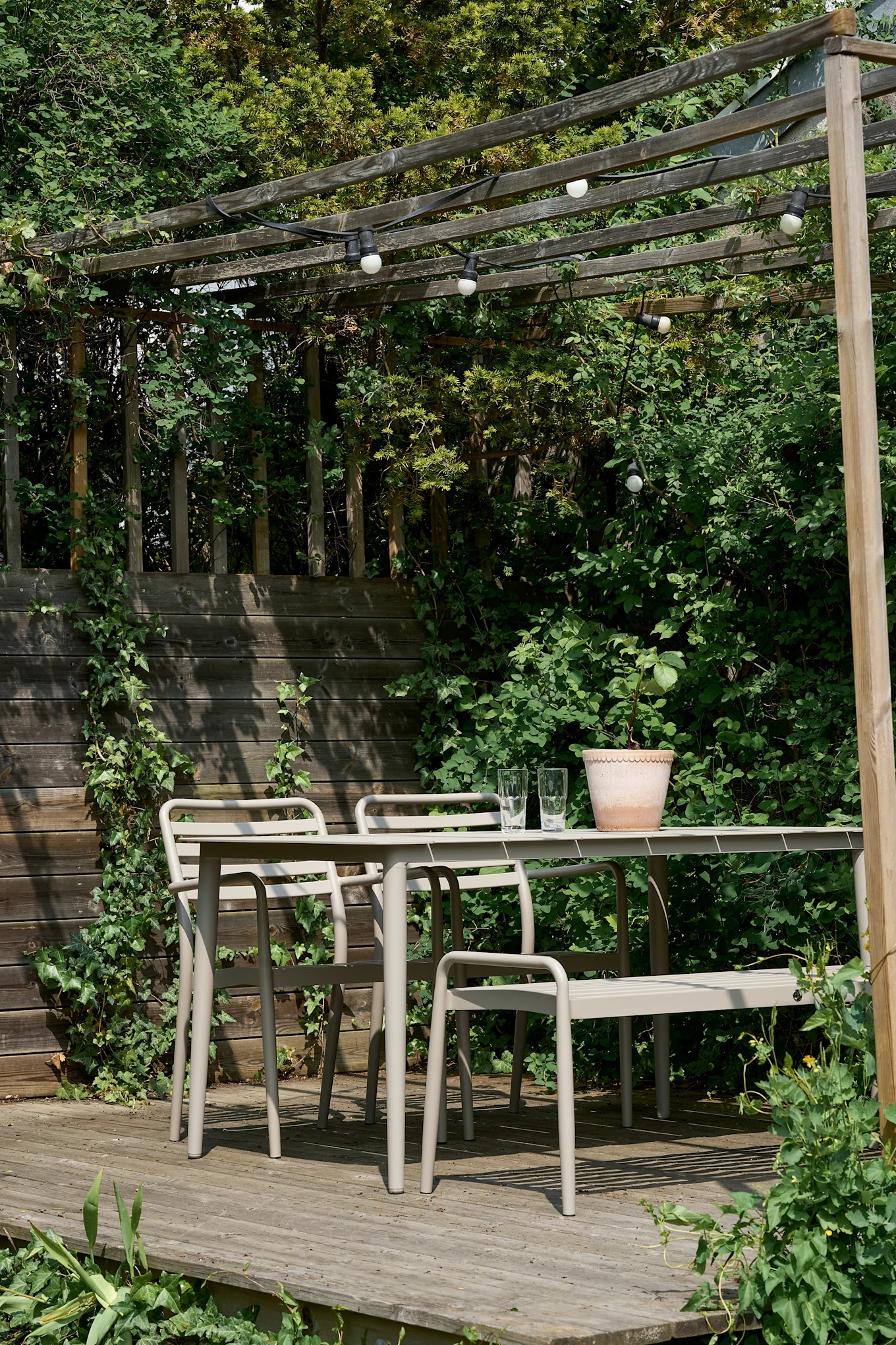 Un ensemble repas extérieur avec table et chaises en métal beige sur une terrasse en bois sous une pergola avec des guirlandes lumineuses.