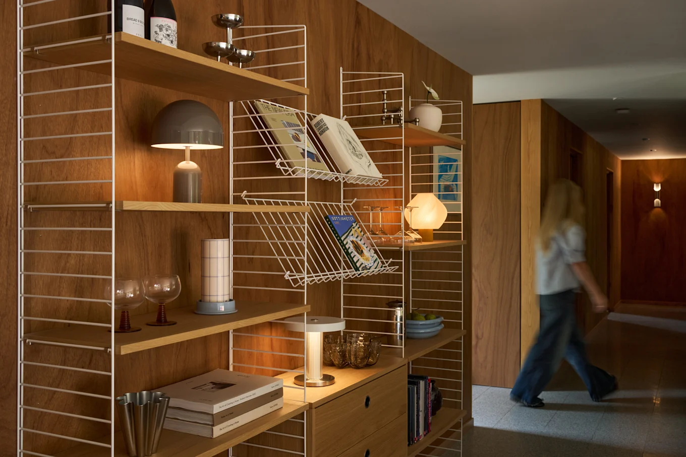 Une personne blonde marche dans un couloir en bois à côté d'une étagère moderne remplie de livres, de lampes et d'objets décoratifs.