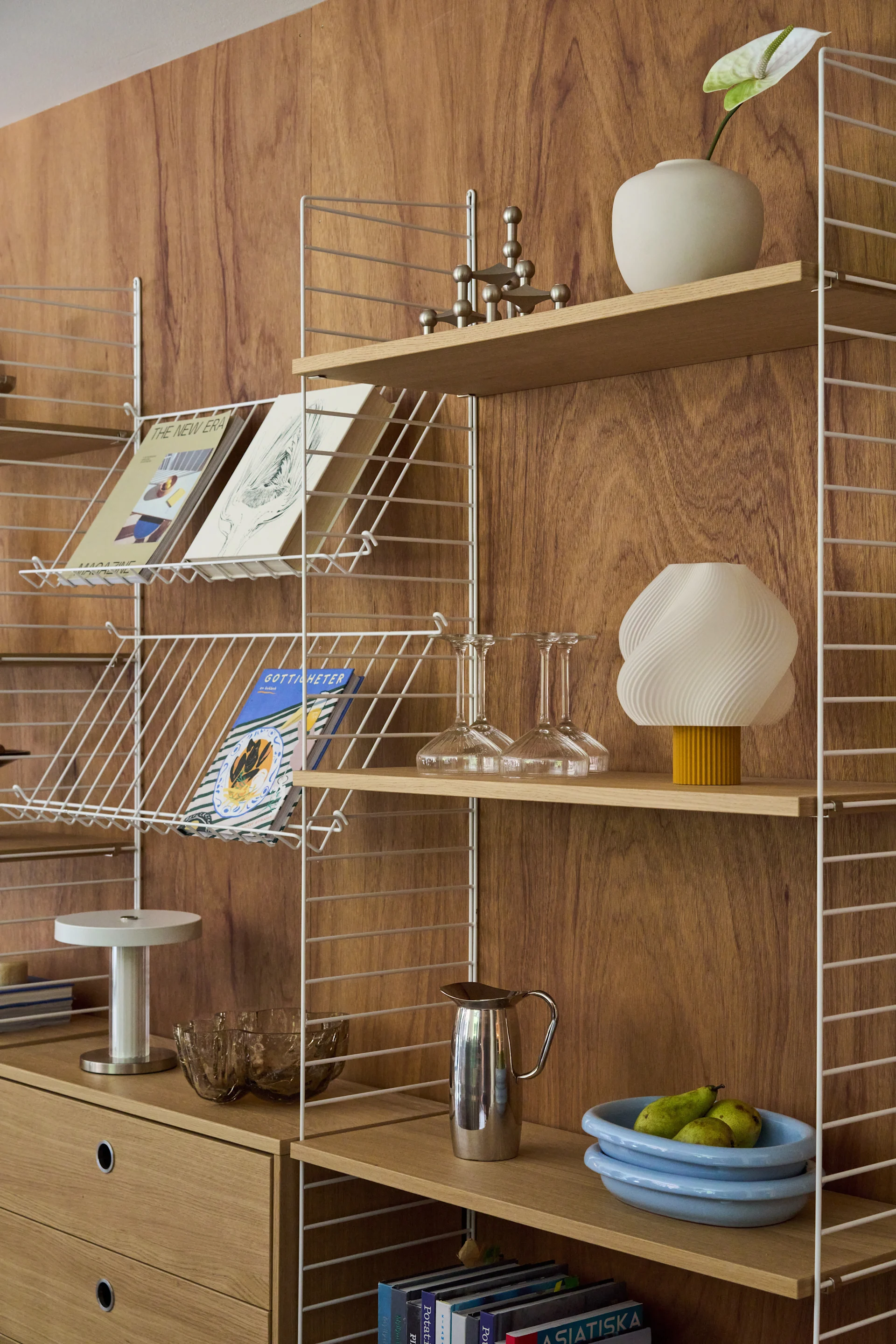 Étagère en bois avec supports en fil blanc, présentant des livres, magazines, un vase blanc avec une fleur, une lampe et un bol de poires.
