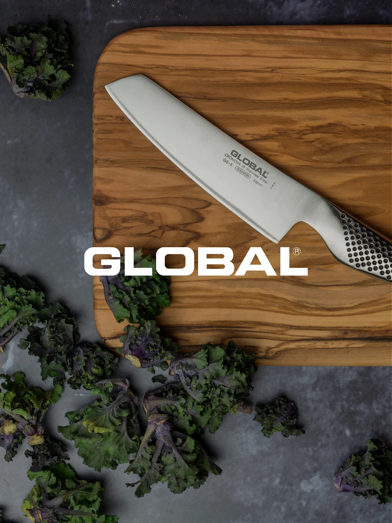 Un couteau Global sur une planche à découper en bois, entouré de légumes-feuilles verts et violets frais.