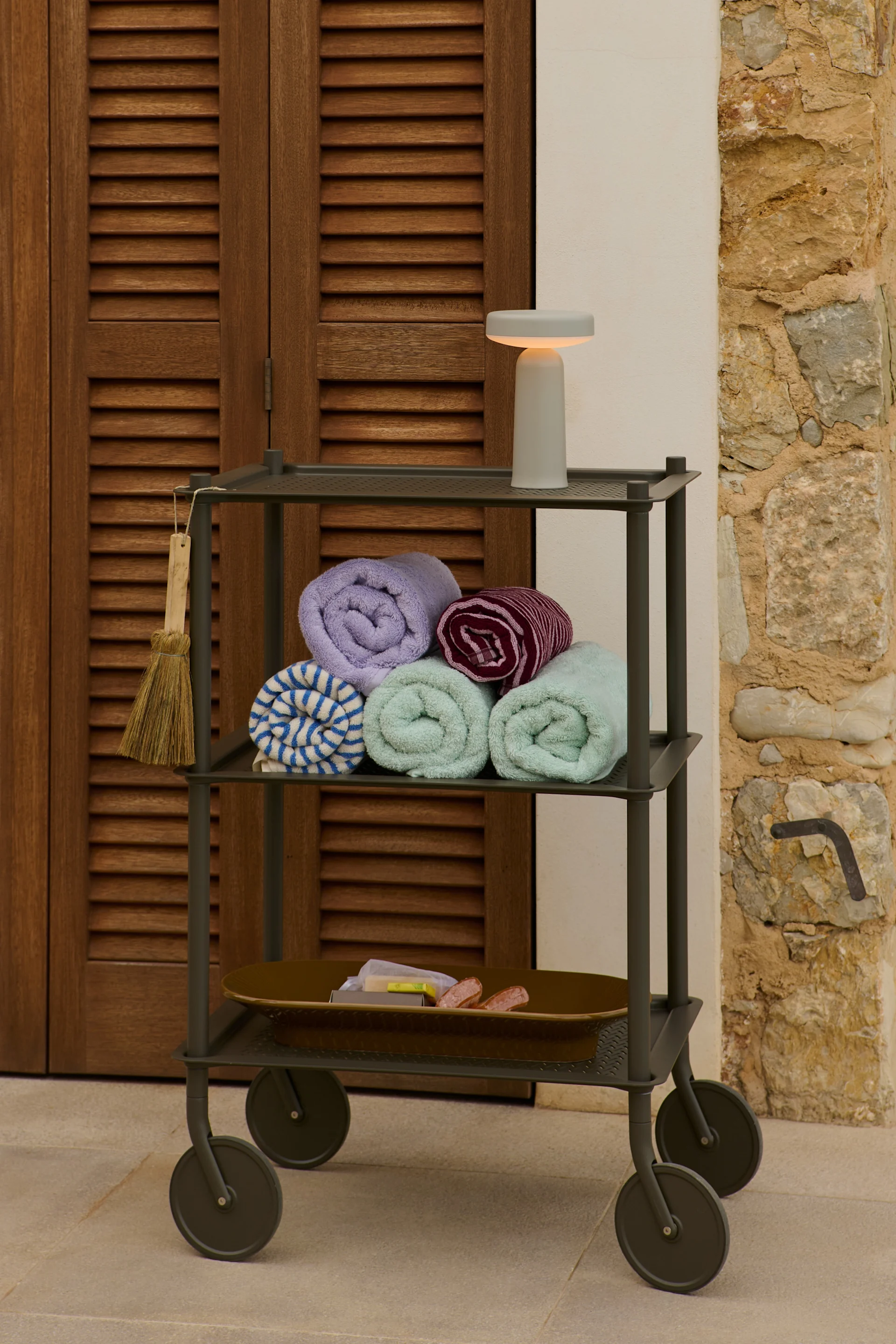 Une desserte à trois niveaux avec des serviettes roulées, des articles de toilette et une lampe à côté d'une porte en bois et d'un mur en pierre.