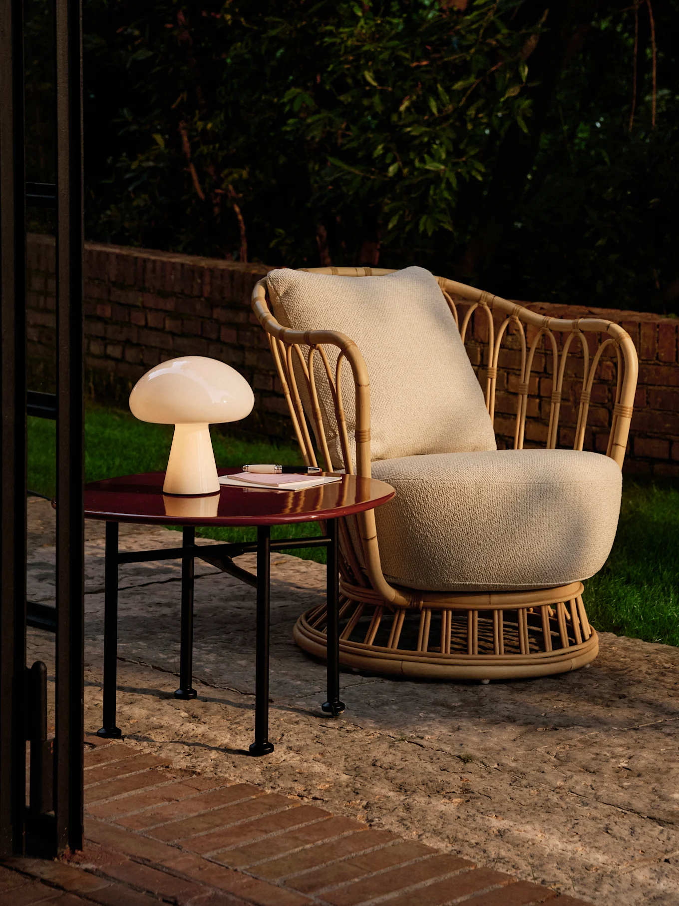Lampe champignon sur une table d'appoint rouge près d'un fauteuil en rotin sur une terrasse au crépuscule.