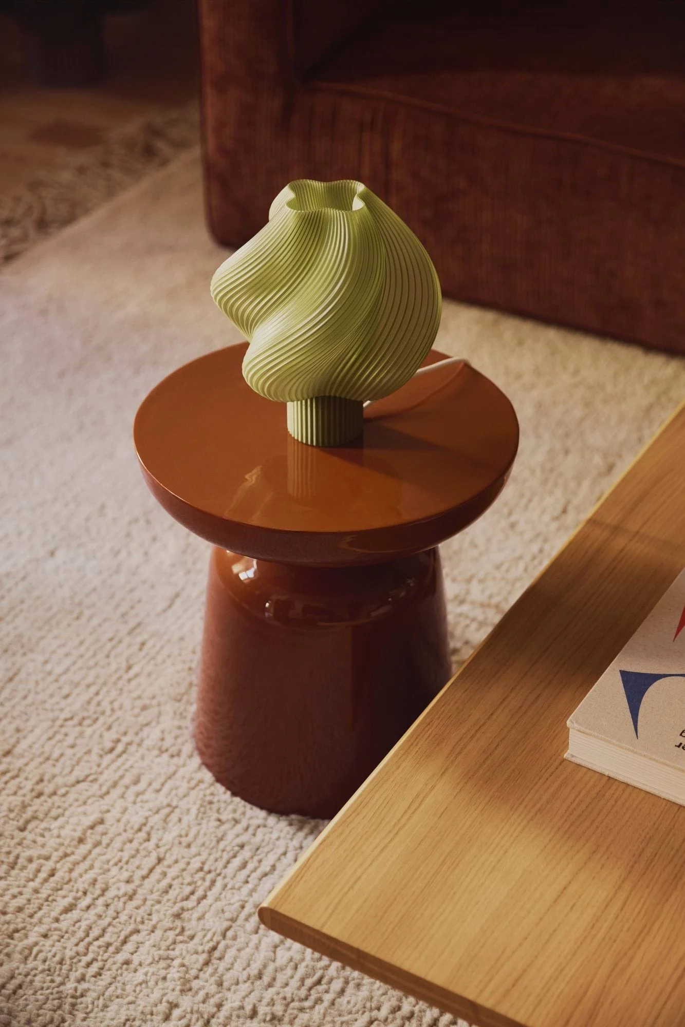 Un vase vert clair strié trône sur une table d'appoint ronde de couleur rouge-brun, posée sur un tapis couleur crème, avec un canapé en arrière-plan.