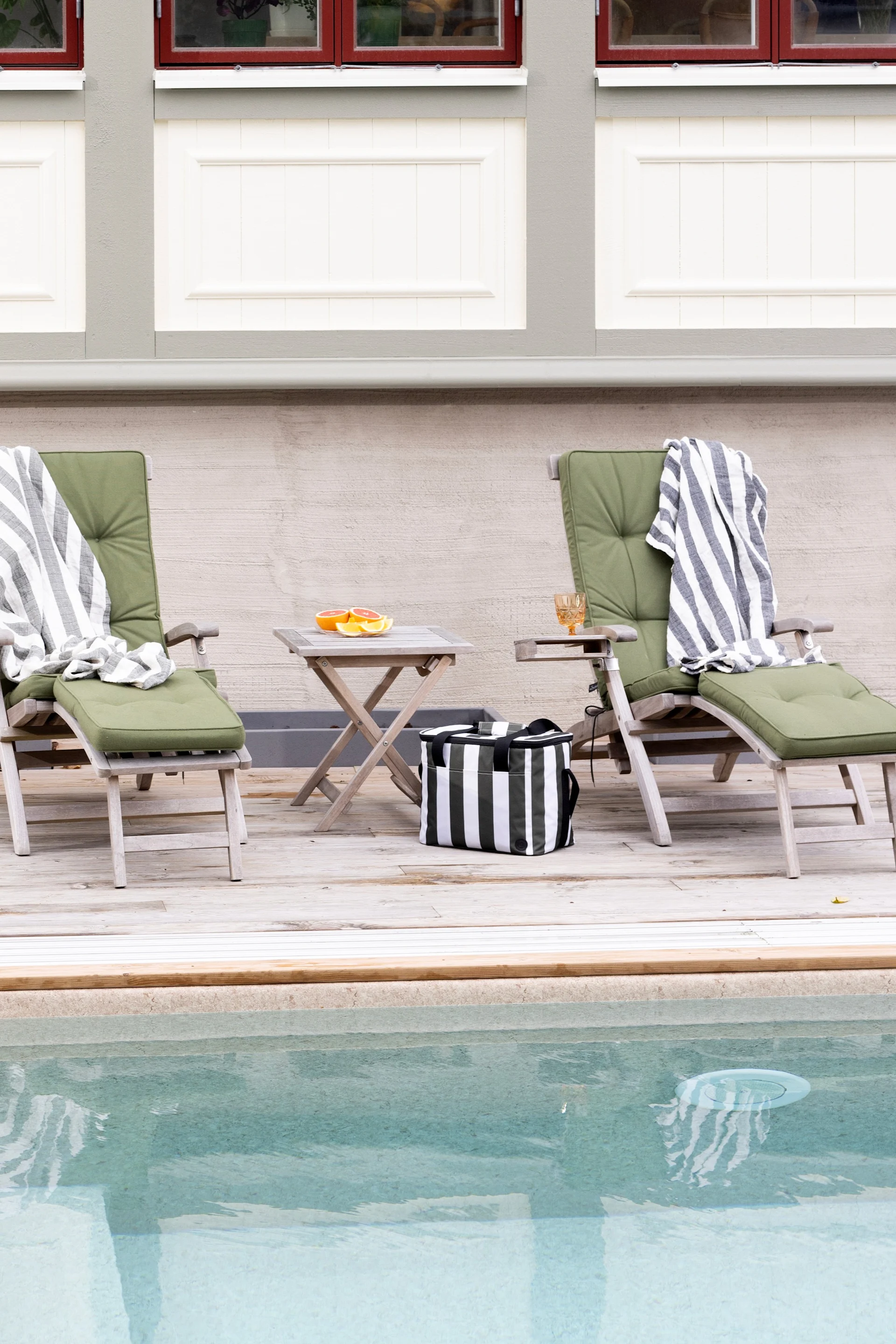 Deux chaises longues en bois avec des coussins verts et des couvertures rayées, posées sur une terrasse en bois au bord d'une piscine. Un sac isotherme et un pamplemousse se trouvent à proximité.