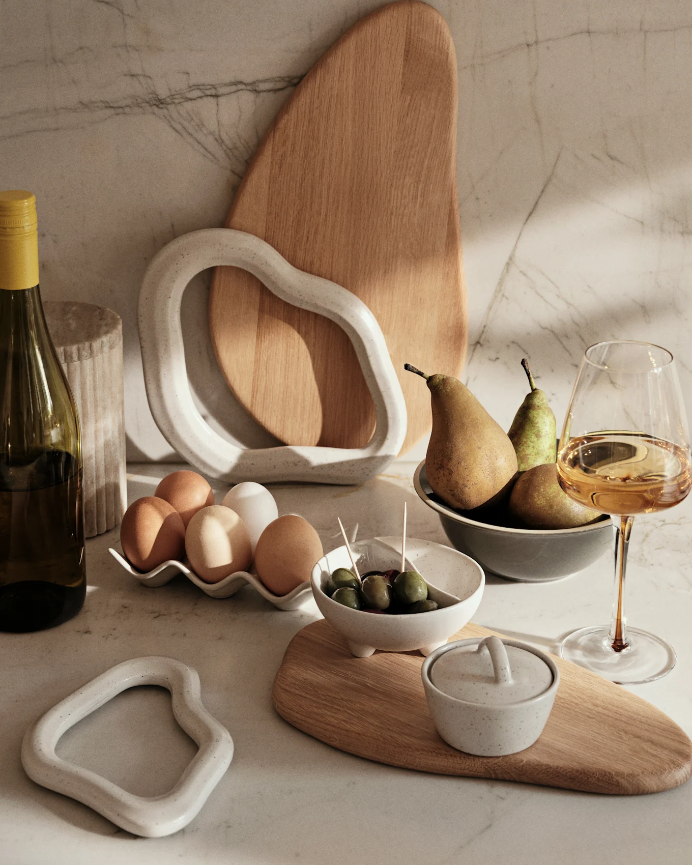Comptoir en marbre avec une bouteille de vin, des œufs, des olives, des poires et des objets décoratifs en céramique et en bois.