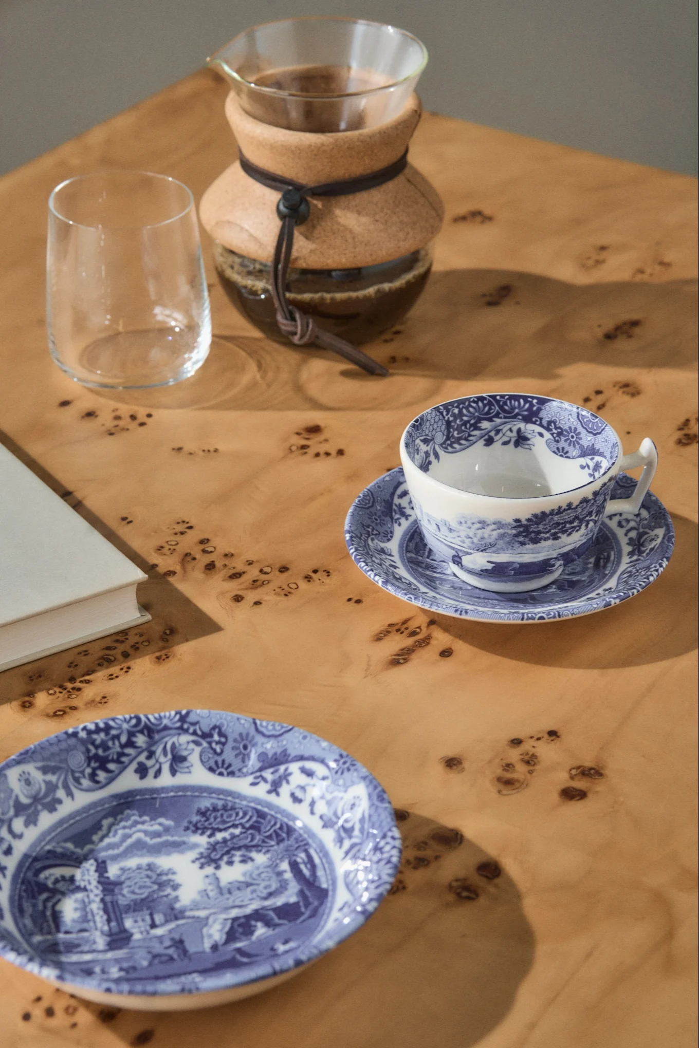 Une cafetière à filtre, un verre vide, une tasse à thé à motifs bleus et blancs sur une soucoupe, et une assiette assortie sur une table en bois clair.