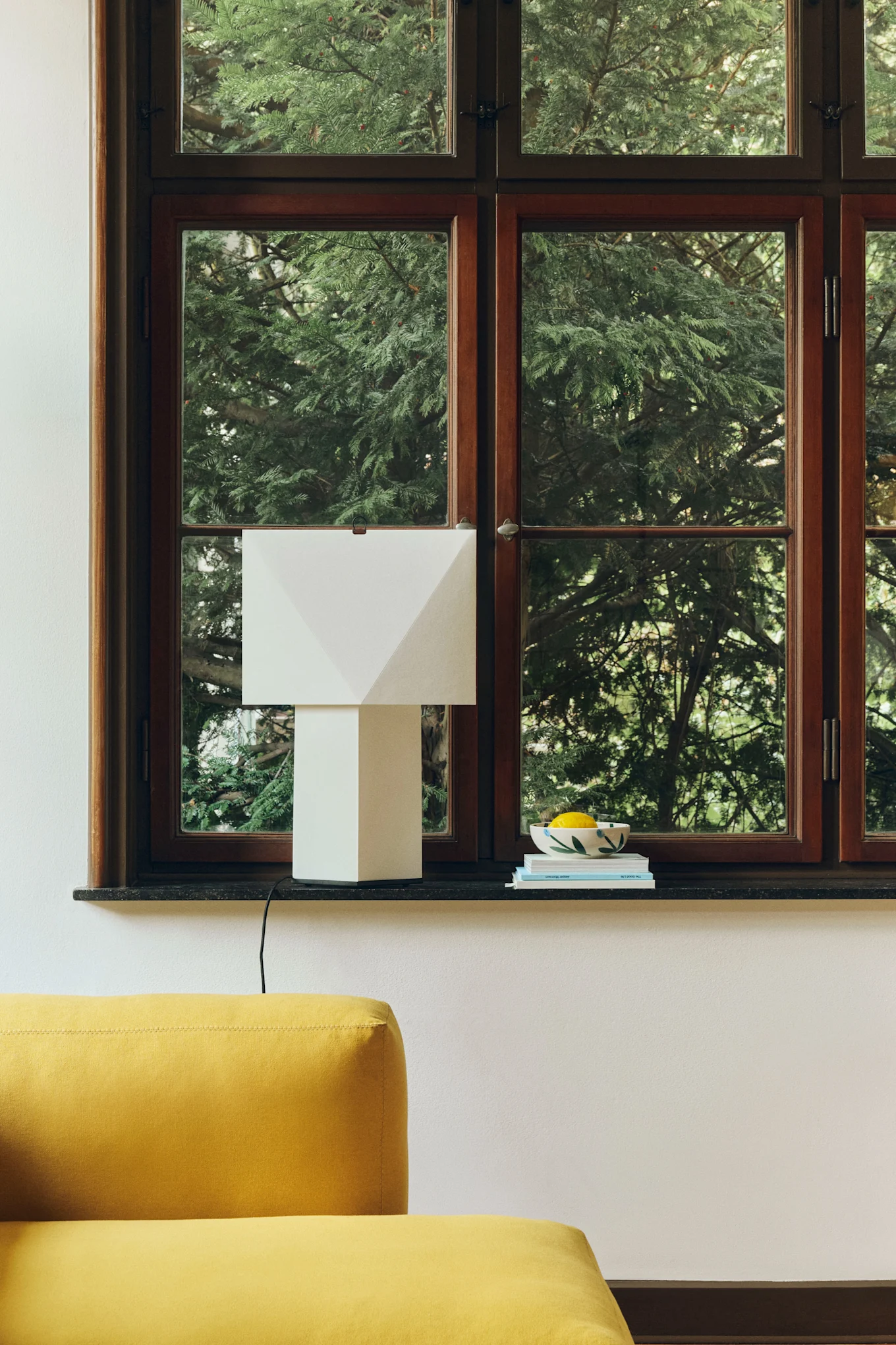 Une lampe de table blanche aux lignes géométriques posée sur un rebord de fenêtre sombre, à côté d'un bol rempli de citrons et de livres, avec un canapé jaune au premier plan.