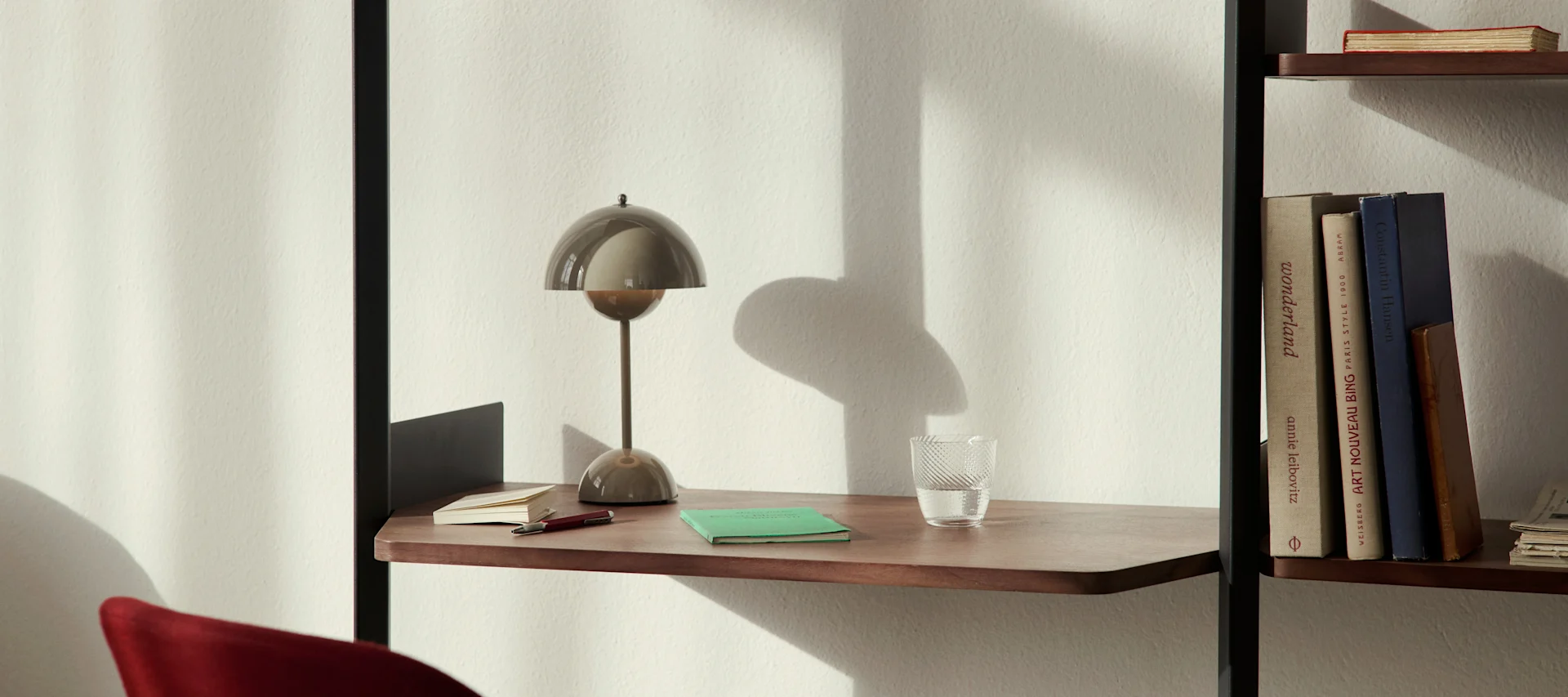 Une lampe de bureau moderne en forme de champignon, un verre d'eau et des cahiers sur une étagère en bois près d'une bibliothèque.