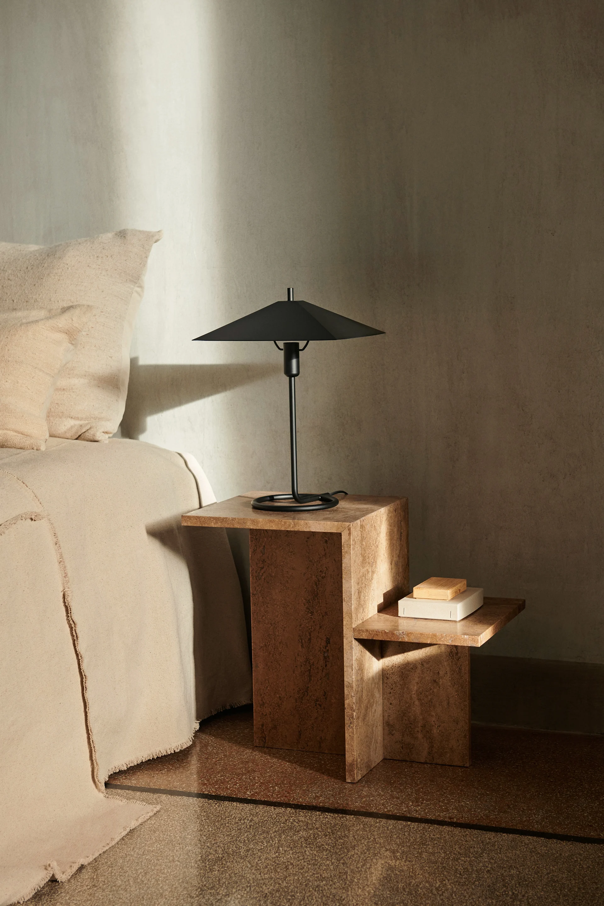 Une scène de chambre à coucher avec un lit beige à côté d'une table de chevet marron texturée à deux niveaux, surmontée d'une lampe de table noire au design moderne.
