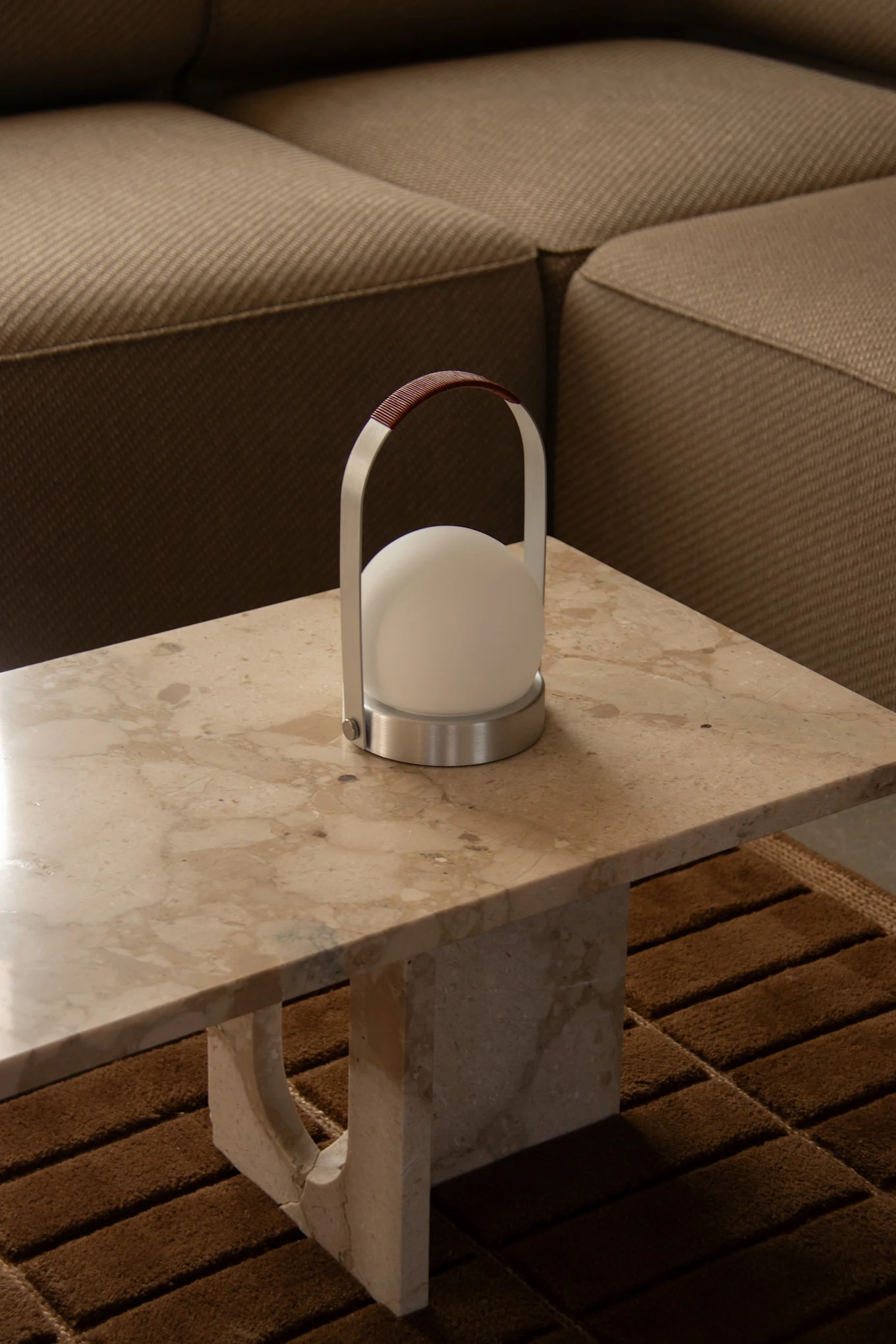Une lampe portable moderne, dotée d'un abat-jour sphérique blanc, d'une structure en métal brossé et d'une poignée en cuir marron, trône sur une table en marbre clair.