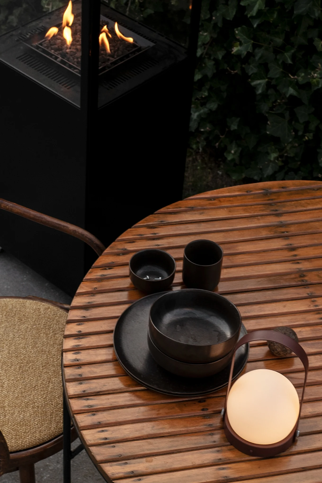 Vous voyez ici la lampe Carrie, également adaptée à une utilisation en extérieur, dans la couleur Burned Red, posée sur une table de jardin en bois, à côté d'un couvert noir. À côté se trouve une chaise et derrière la table, un brasero crépite.