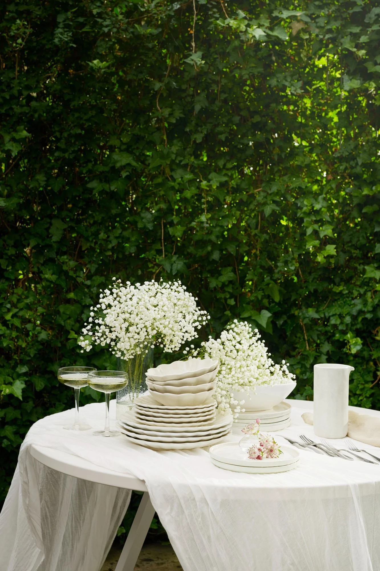 Table d'extérieur dressée avec vaisselle blanche, fleurs de gypsophile, flûtes à champagne et couverts sur une nappe blanche, devant un feuillage vert luxuriant.