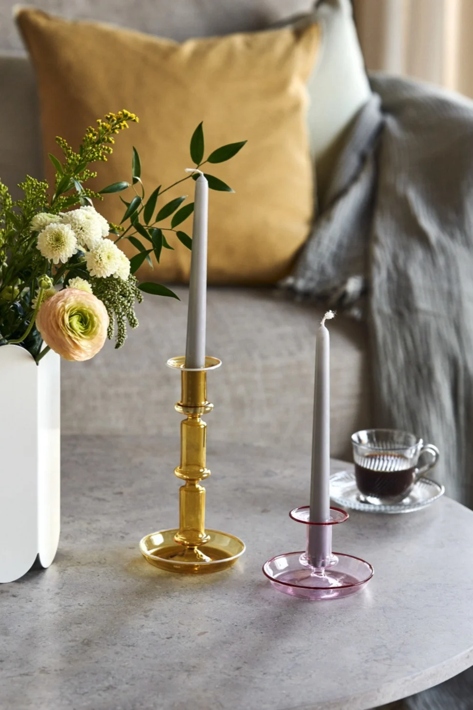 Deux bougeoirs en verre (ambre et rose) avec des bougies grises sur une table, près d'un vase de fleurs et d'une tasse de café.