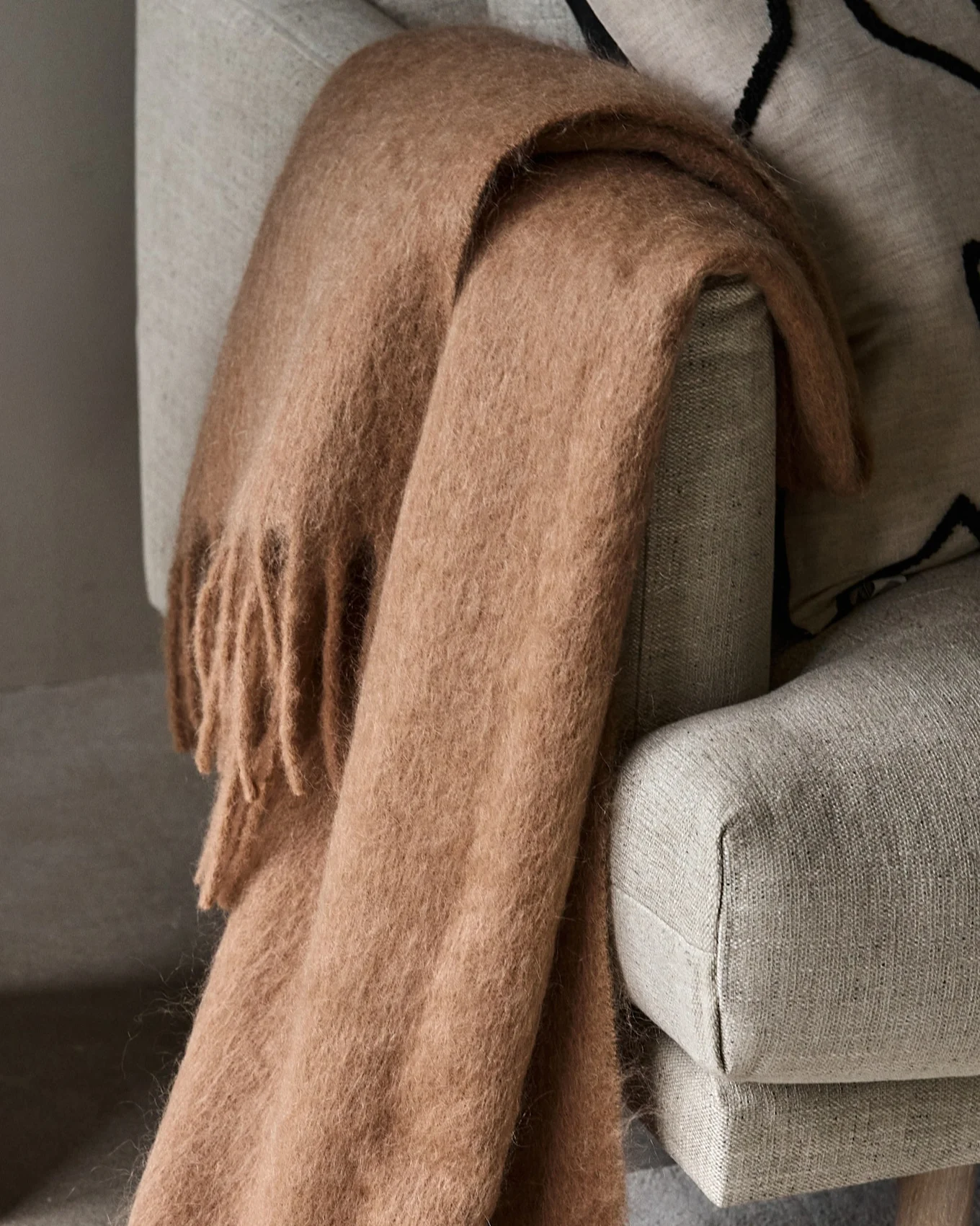 Plaid douillet marron clair et moelleux avec des franges, drapé sur un fauteuil gris clair.