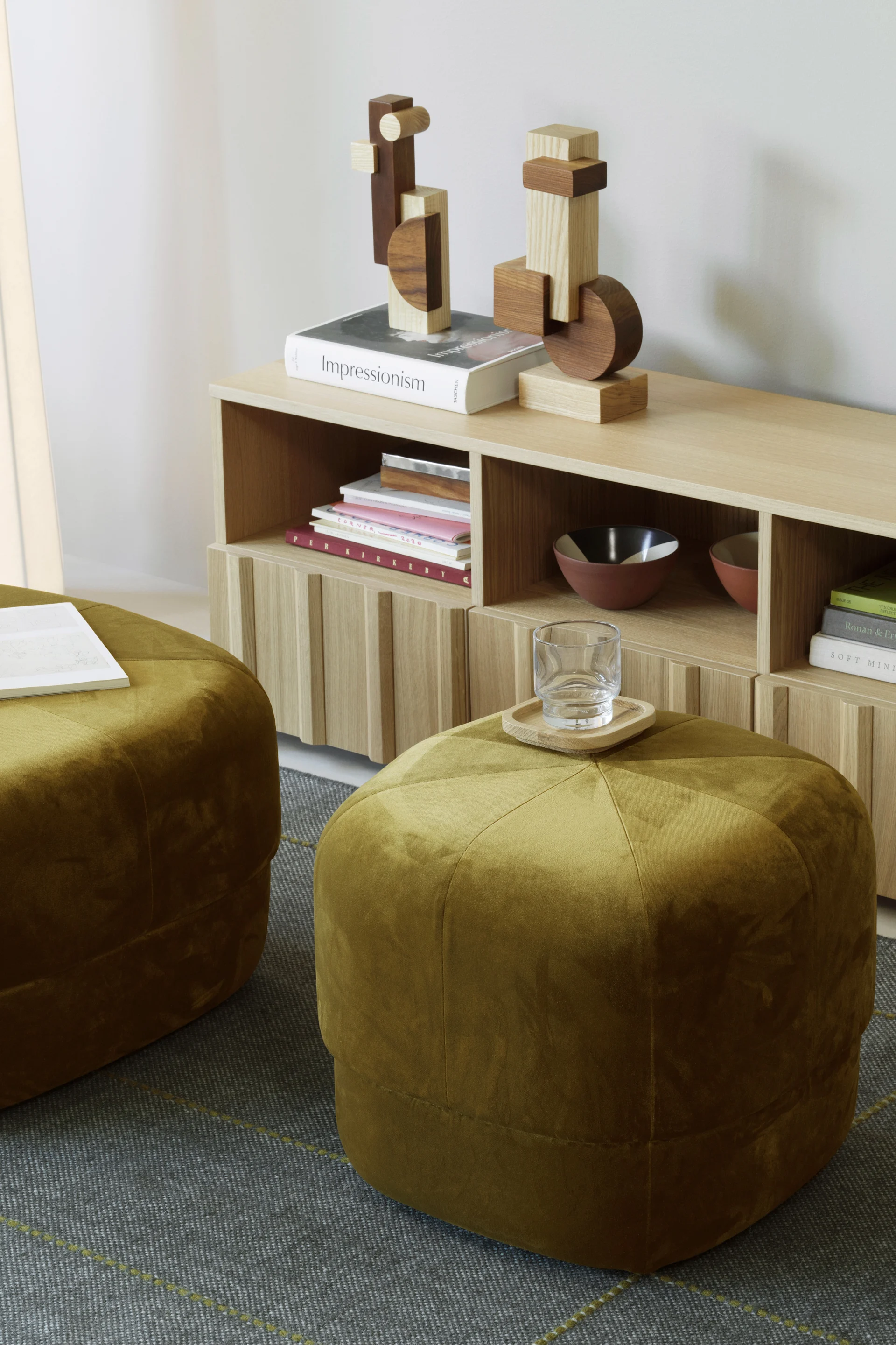 Deux poufs en velours vert olive posés sur un tapis à motifs, à côté d'une étagère en bois clair garnie de livres et de décorations en bois.