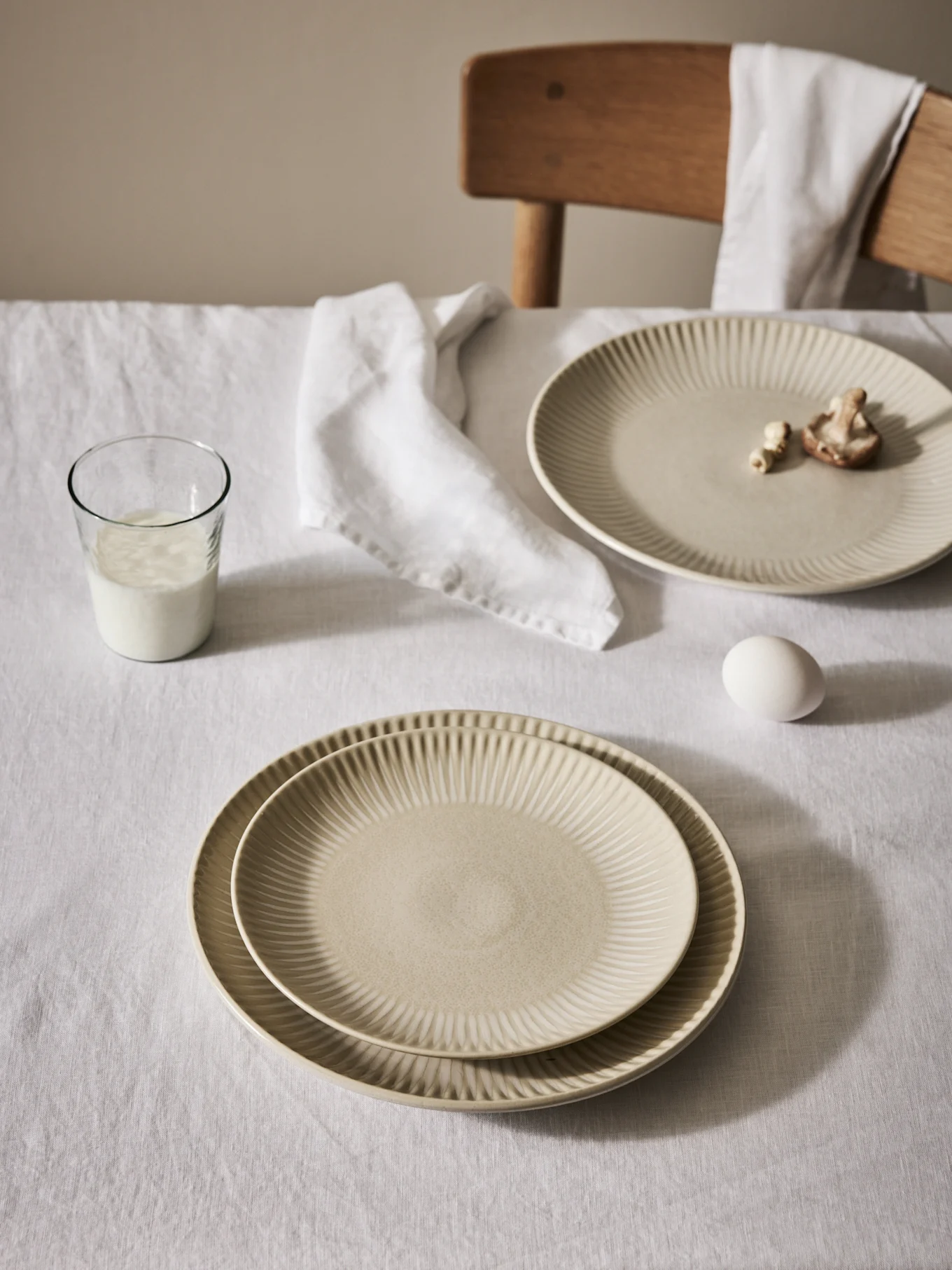 Table dressée avec des assiettes beiges nervurées, un verre de lait, un œuf et une serviette sur une nappe blanche.
