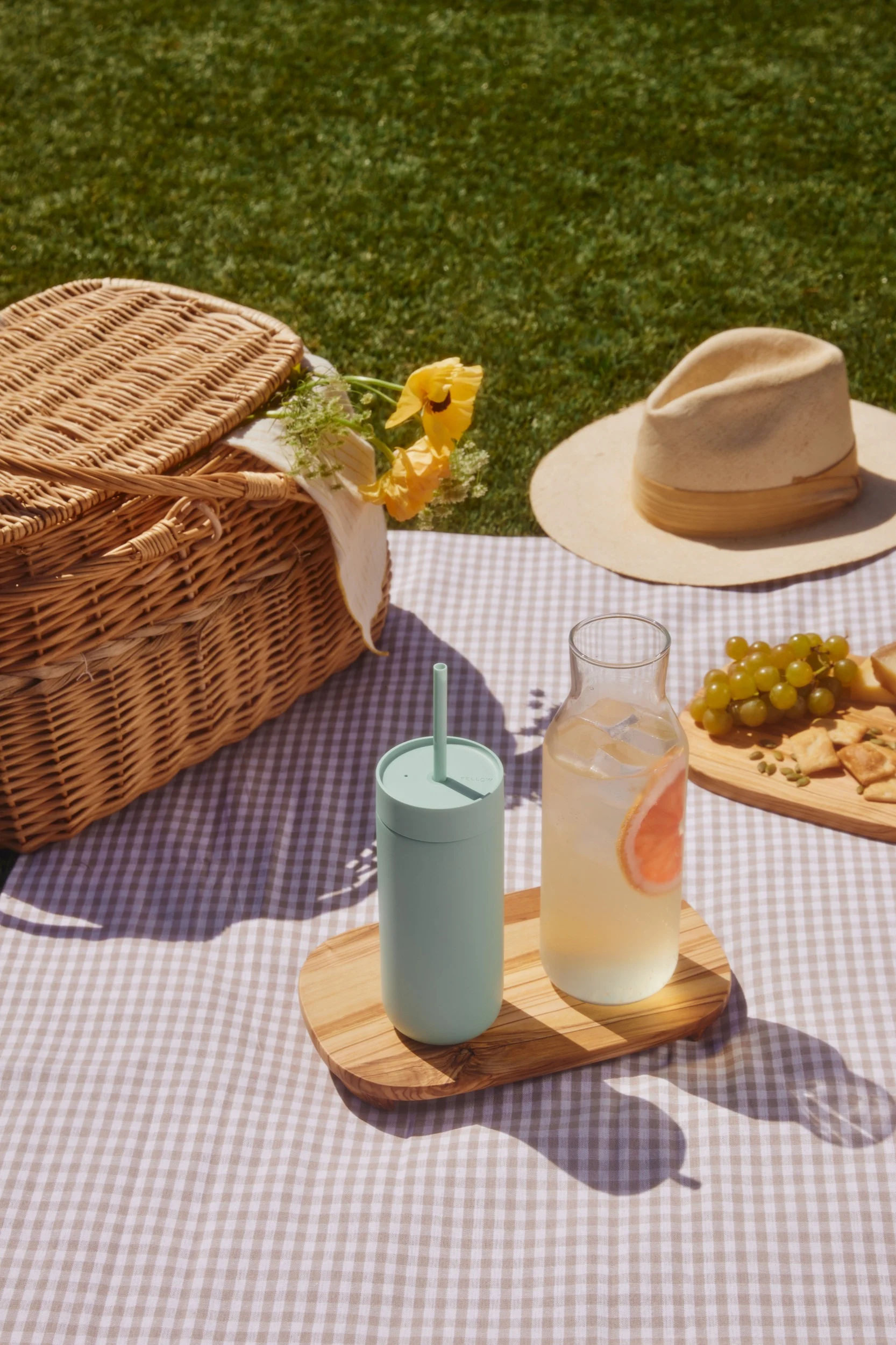 Pique-nique sur une nappe à carreaux: panier en osier, gobelet vert clair, boisson au pamplemousse, raisins et crackers sur l'herbe.